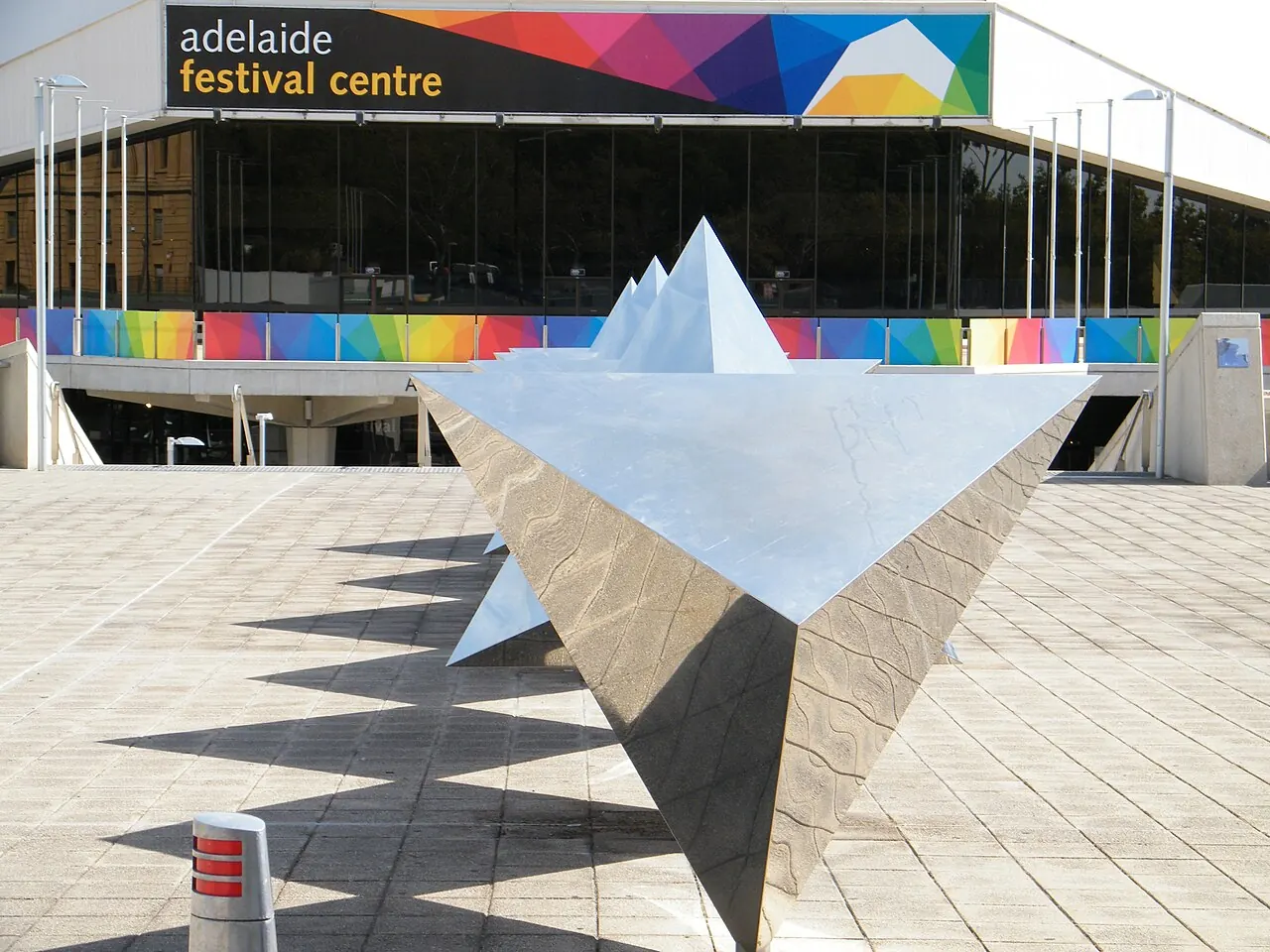 A silver triangular sculpture on the plaza with a colourful sign
