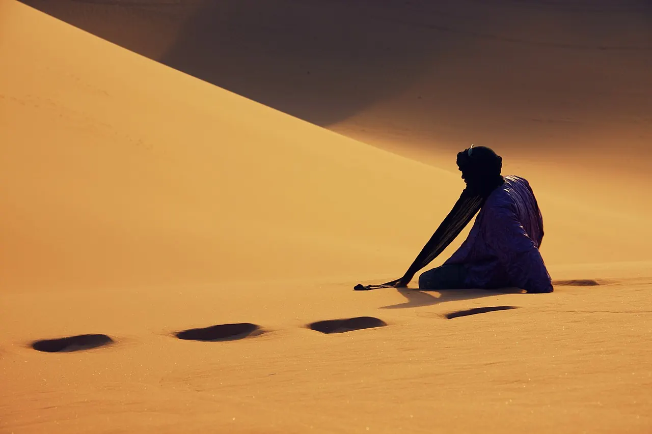 A figure sitting on a sand dune with footprints in the sand