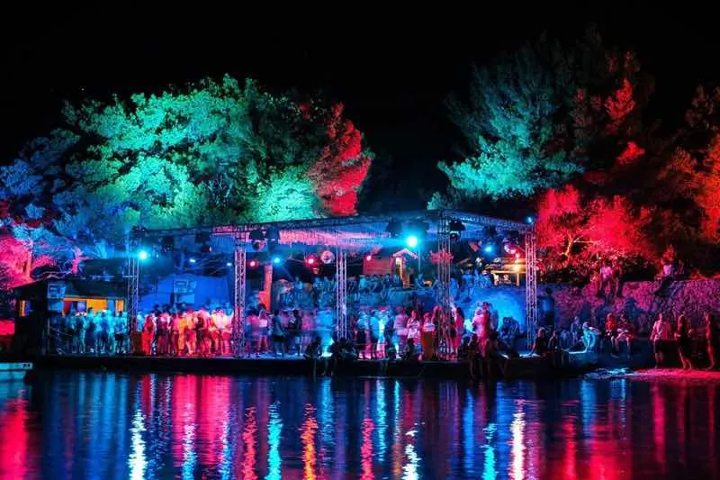 A lit stage beside the water among the trees at The Garden, Tisno, at Dimensions