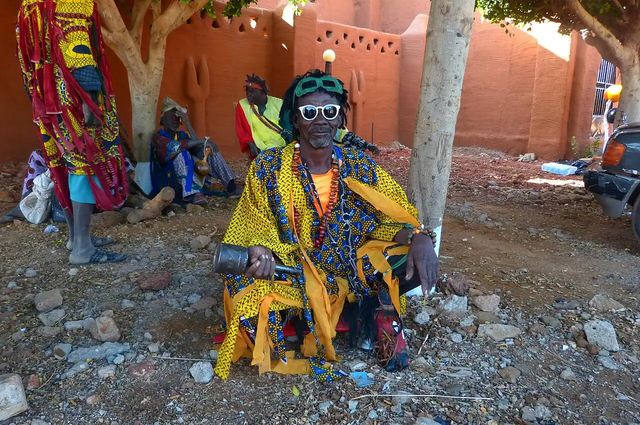A person in a bright yellow costume and sunglasses seated in front of a red wall