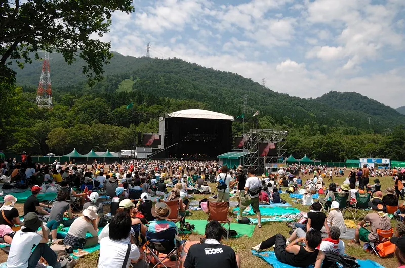 A huge festival crowd on grass in front of a stage among mountains