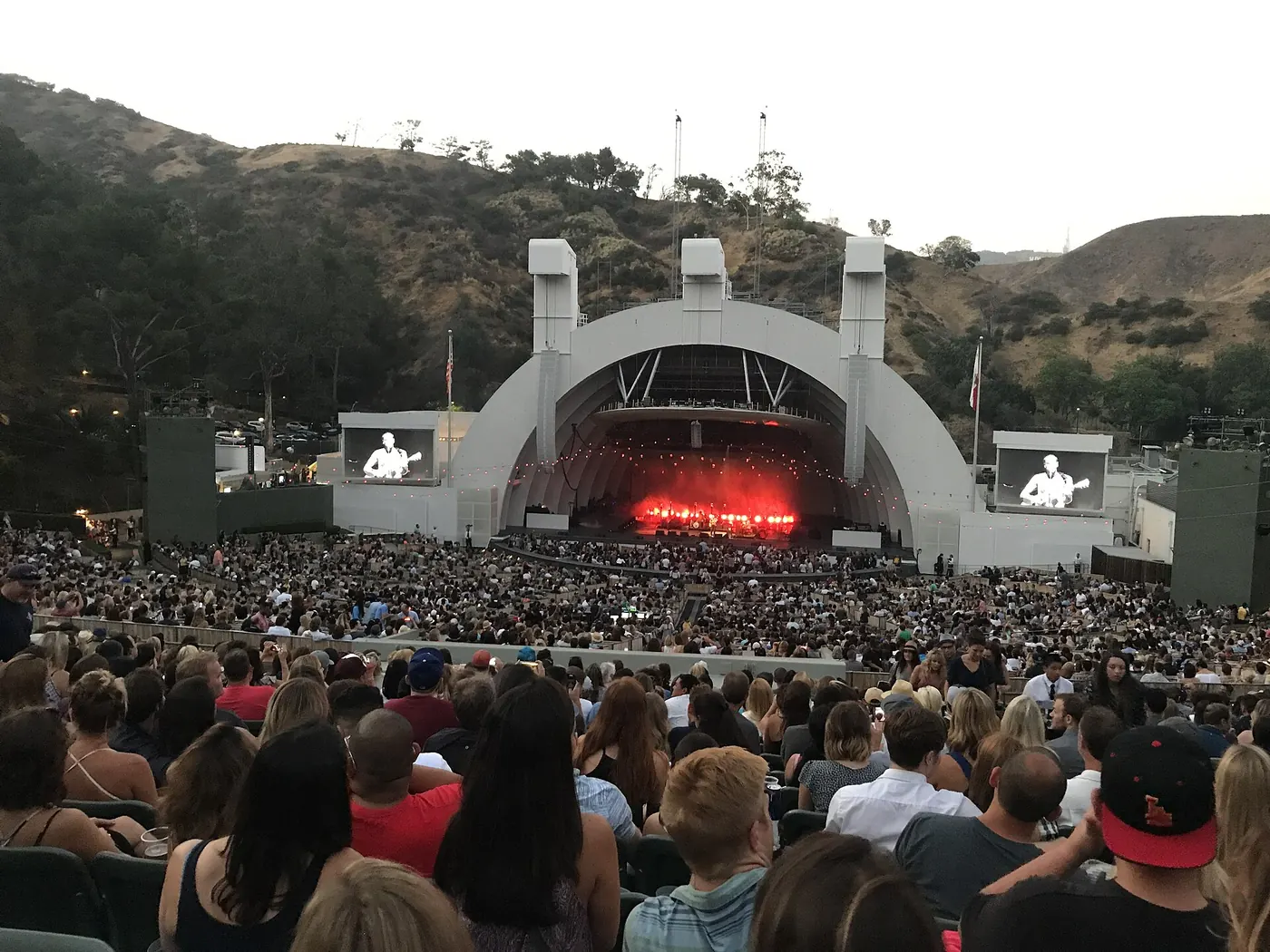 A packed hillside audience at dusk facing the white Hollywood Bowl shell, with video screens and red stage light