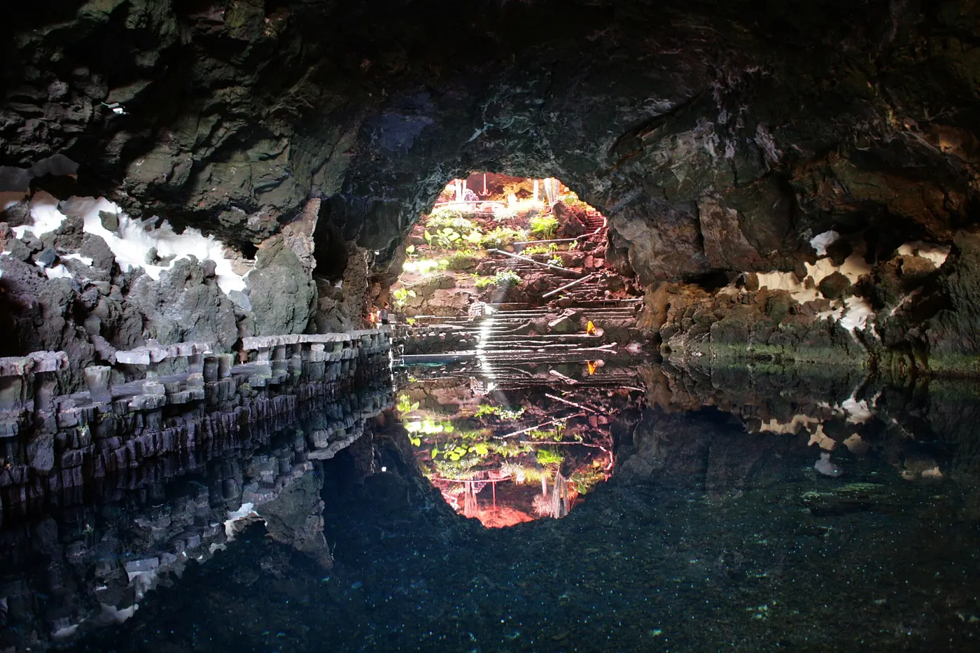 Still dark water inside a lava tube mirrors a bright arched opening with plants and steps above, framed by rough volcanic rock