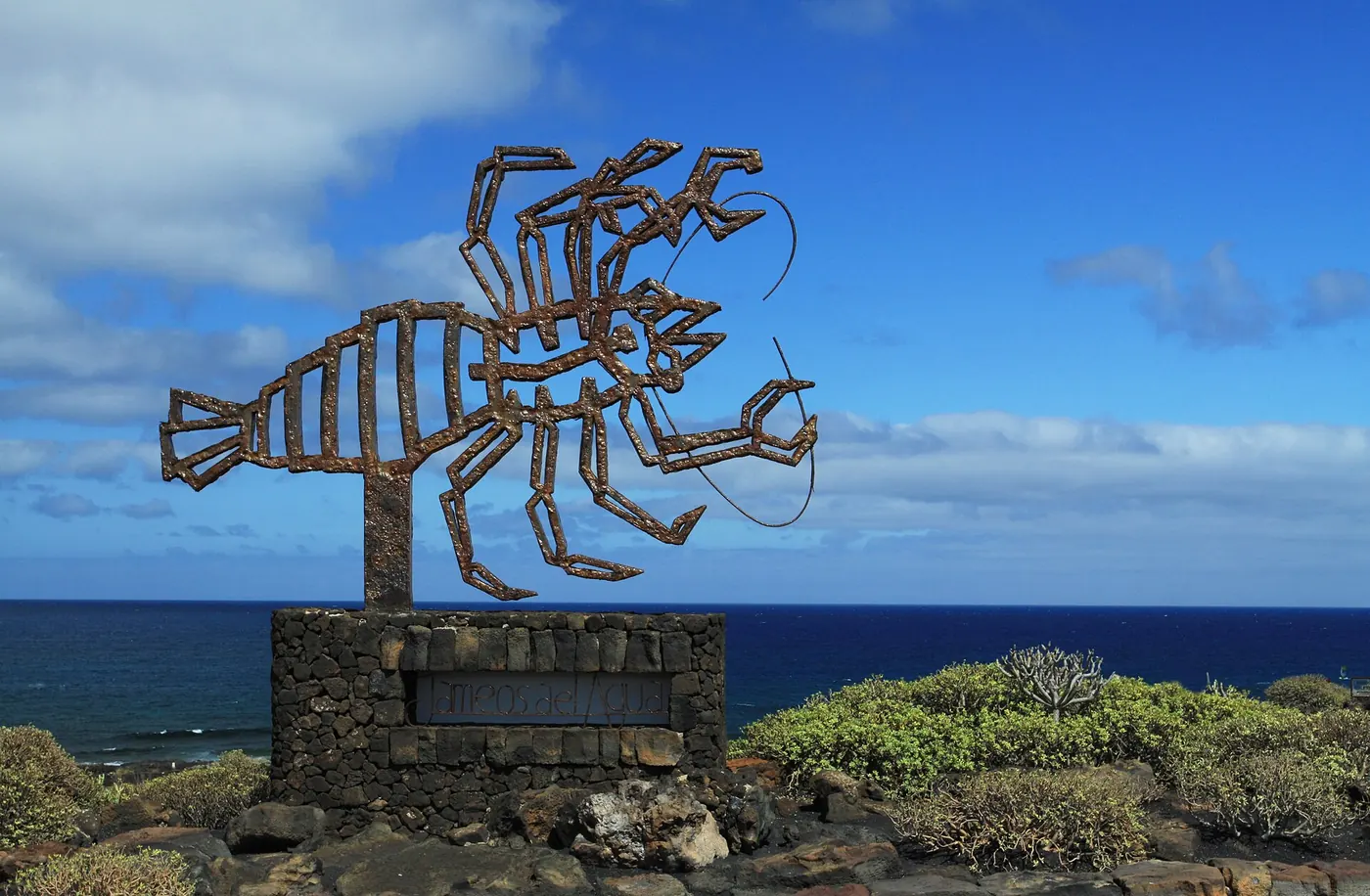 Rusted metal crustacean sculpture on a lava stone plinth marking the Jameos del Agua entrance, with the Atlantic behind