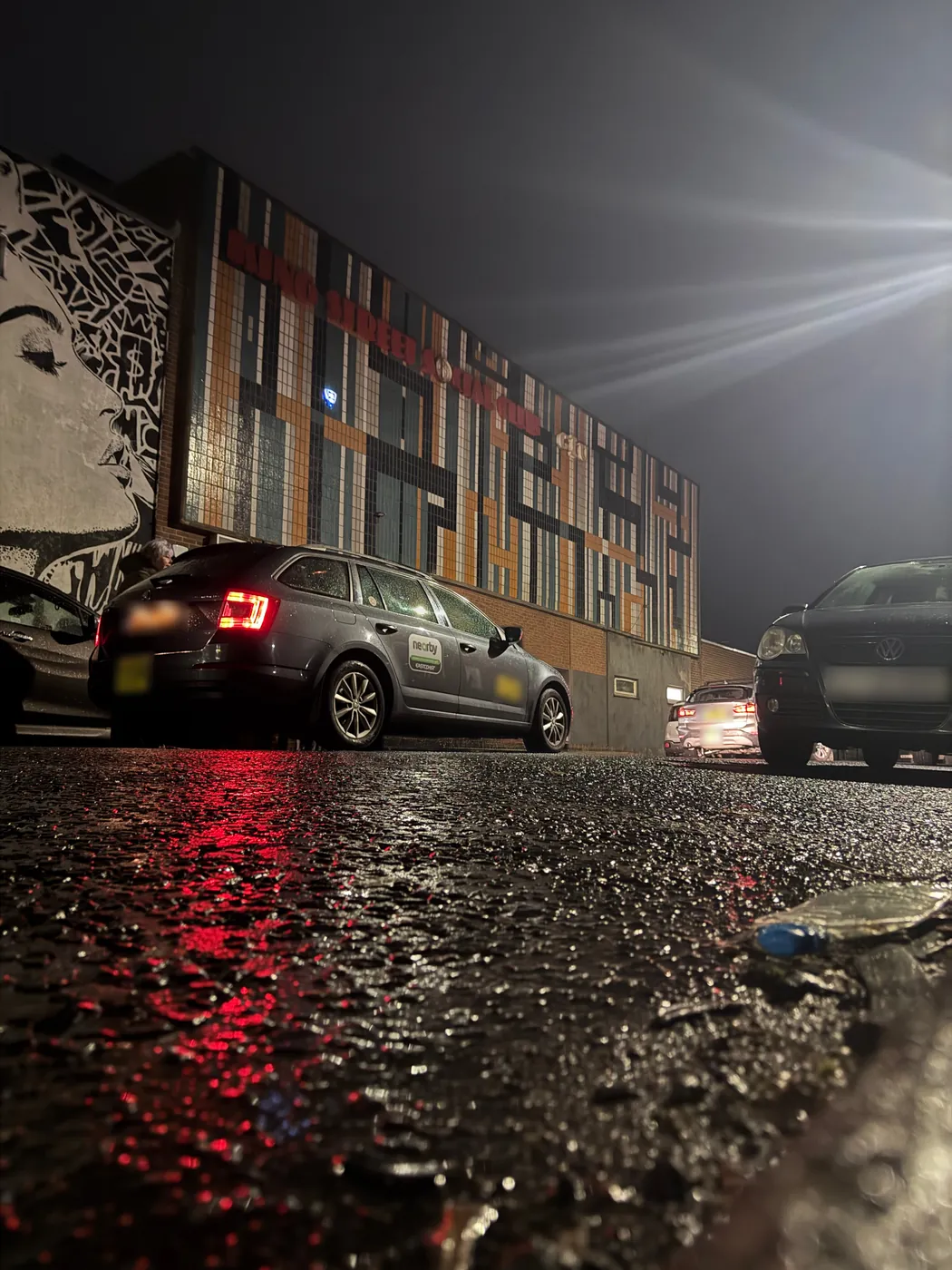 King Street Social Club's tiled facade at night seen from road level, red tail-light reflections on wet tarmac and the mural of a face on the side wall