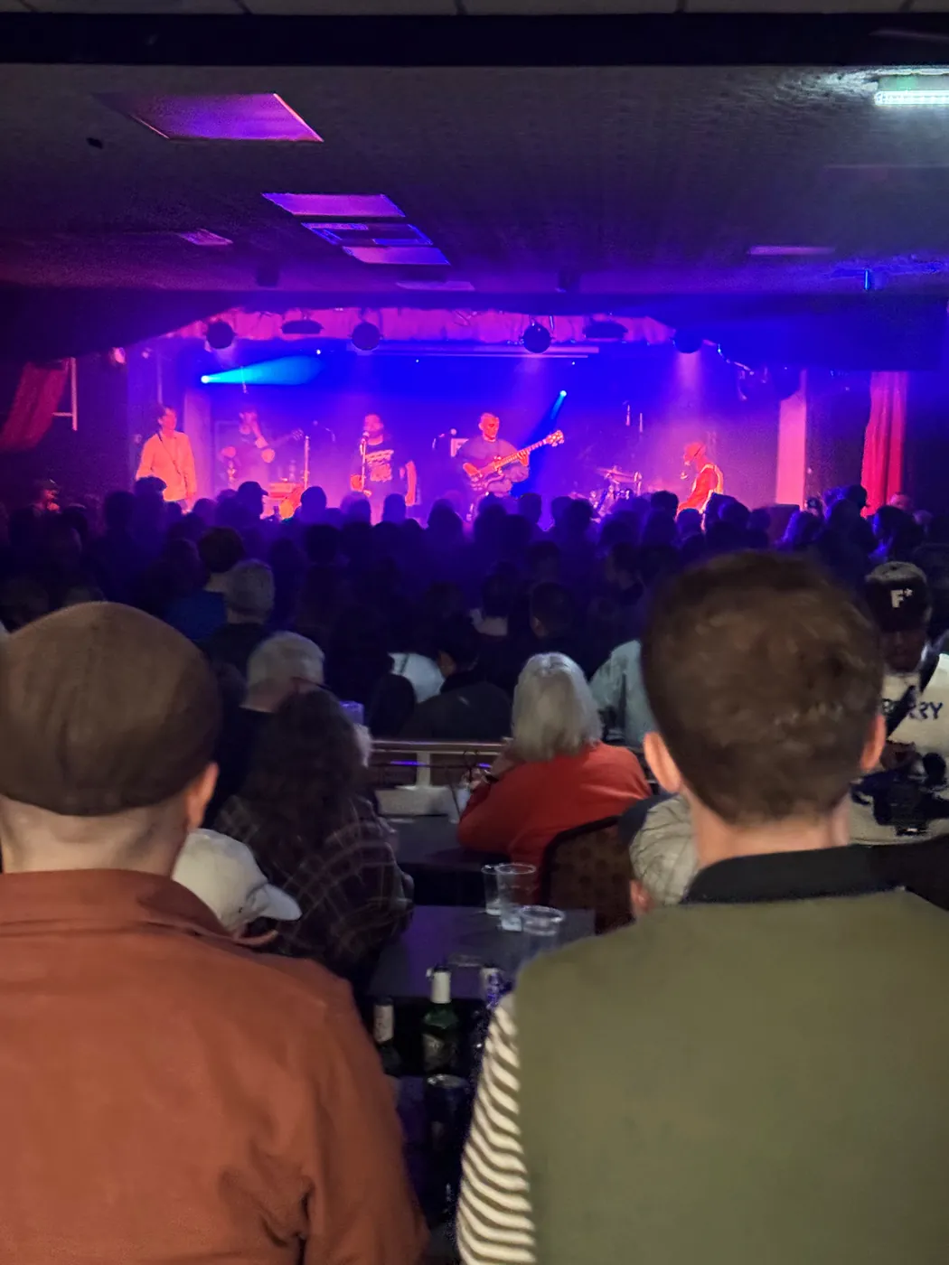 The concert room from the back by the bar: tables, a crowd and the stage lit blue and pink beneath a ruched curtain pelmet