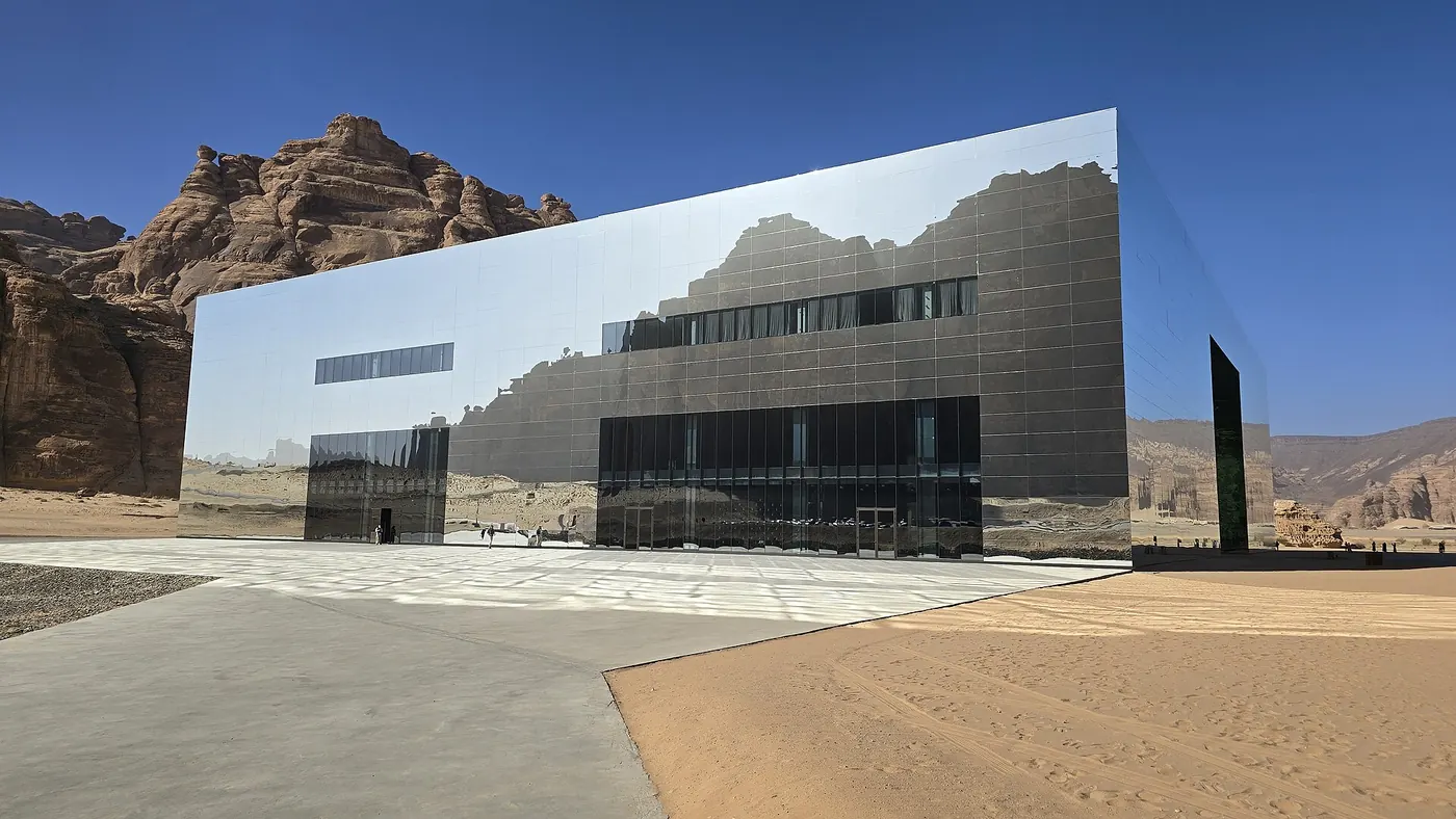 Maraya's mirrored cuboid reflecting AlUla's sandstone cliffs and blue sky, with a paved forecourt and desert sand in front