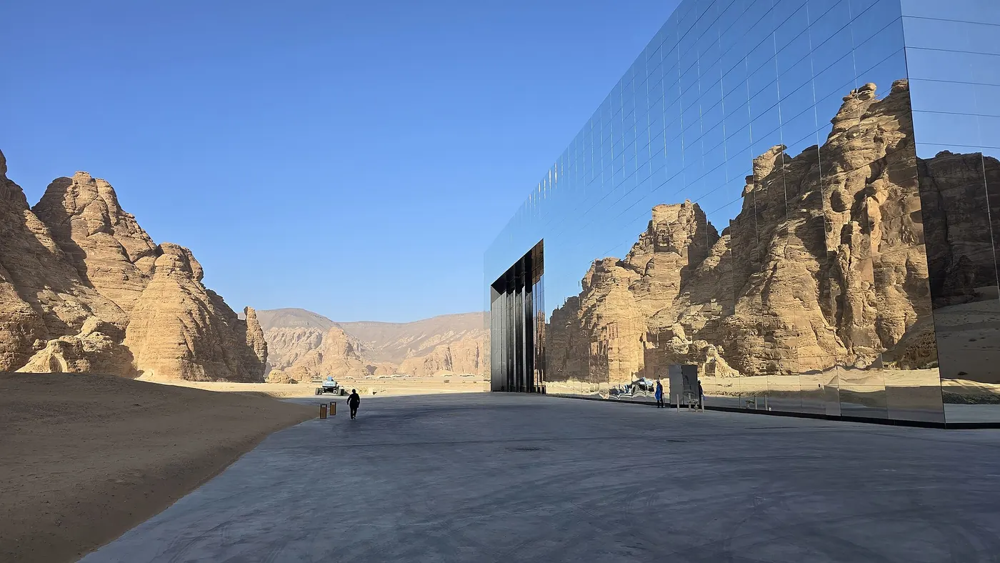 Maraya's tall mirrored wall on a wide paved forecourt, reflecting sandstone crags, with a lone walker between the rocks and the glass
