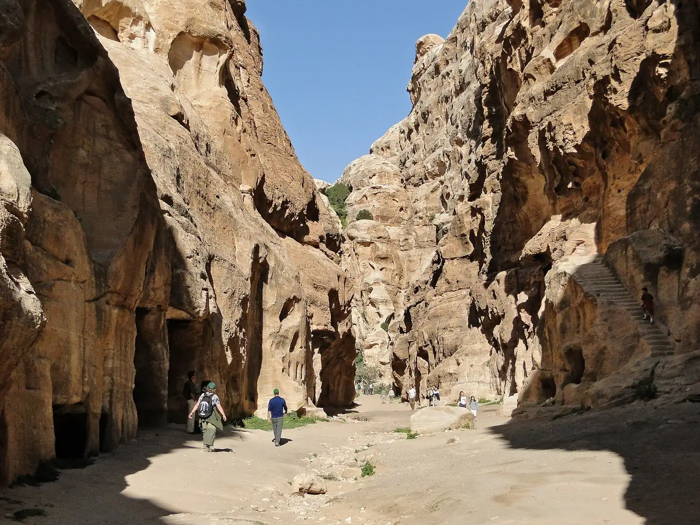 Visitors walking the sandy floor of a narrow sandstone canyon at Little Petra, with a rock-cut stairway climbing the right-hand wall