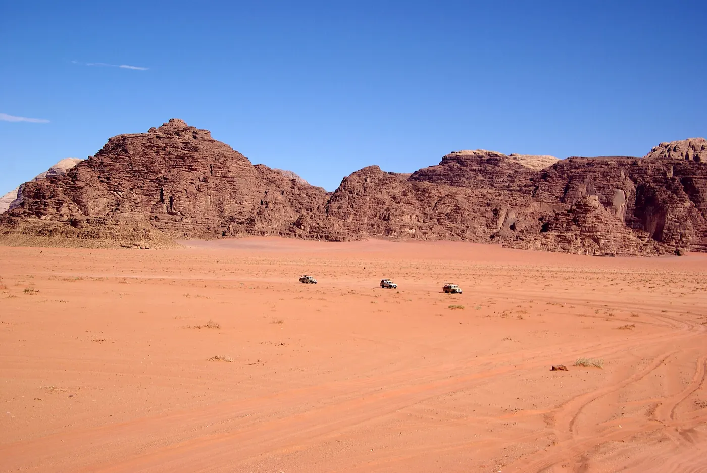 Three jeeps crossing the flat orange sand of Wadi Rum below a long ridge of dark sandstone under a clear blue sky