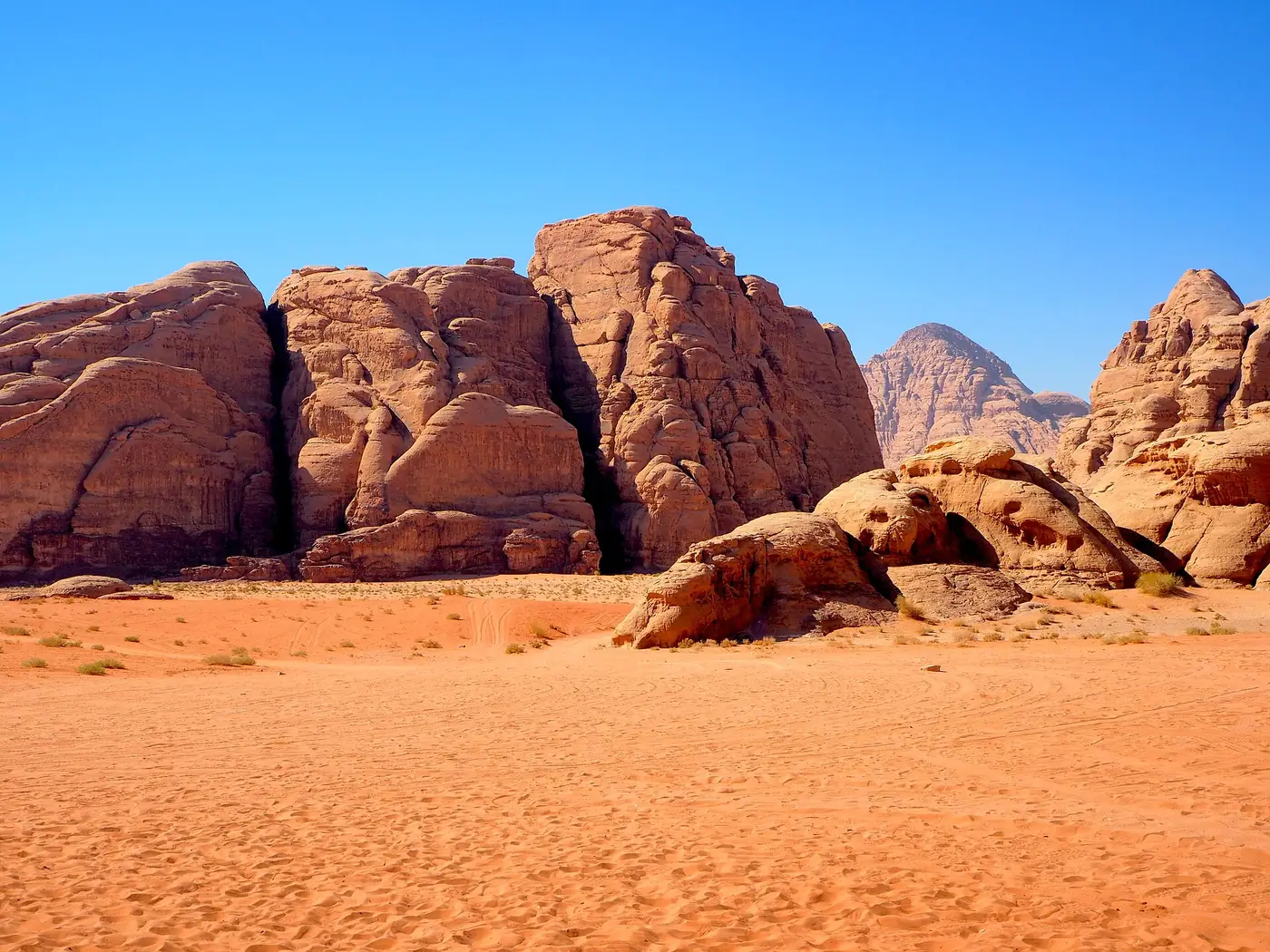 Sun-baked sandstone towers and boulders rising from orange sand in Wadi Rum, with a pointed peak behind and deep blue sky above