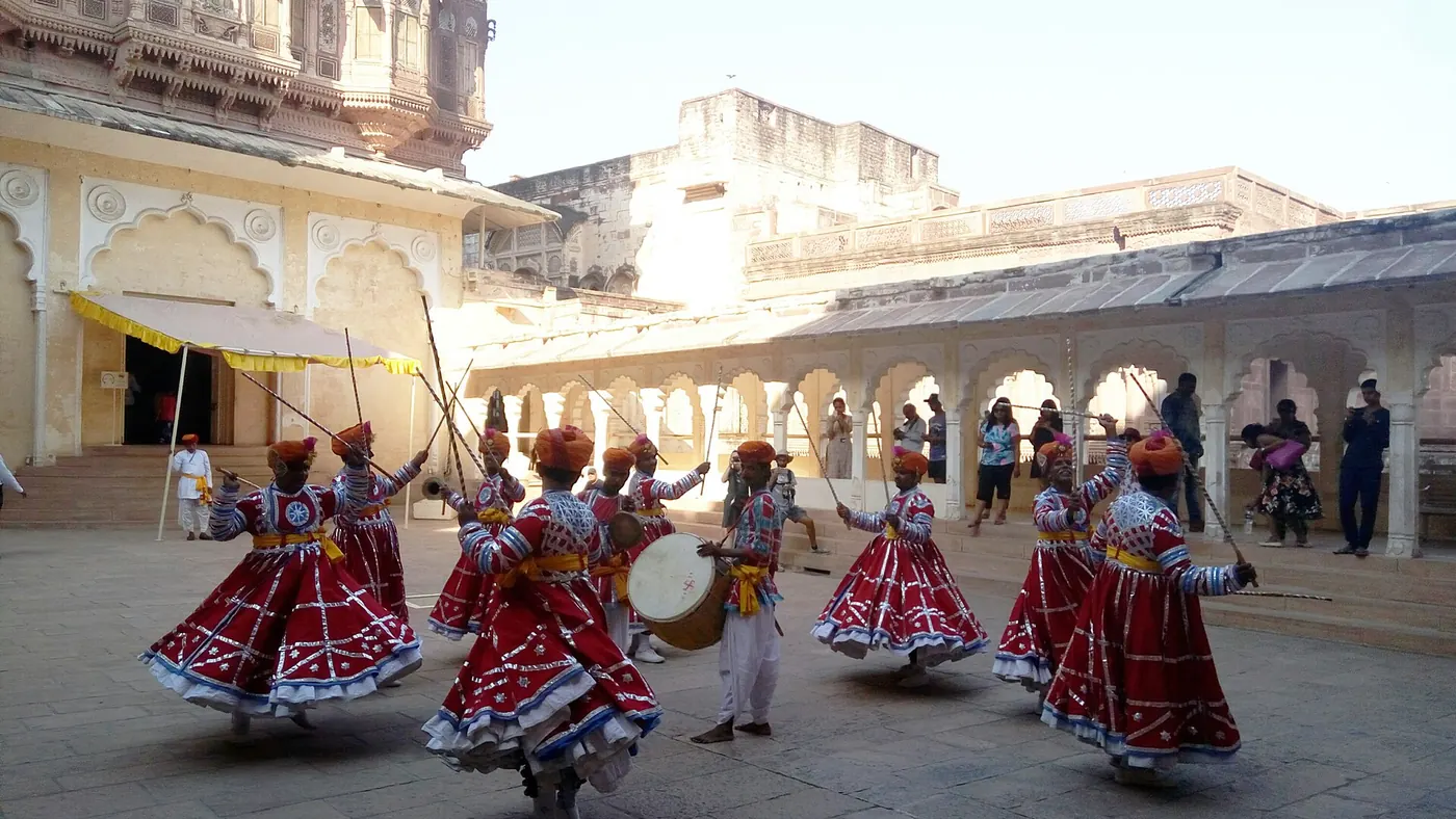 Dancers in red flared costumes carrying sticks and a drummer performing in a sunlit courtyard inside Mehrangarh Fort