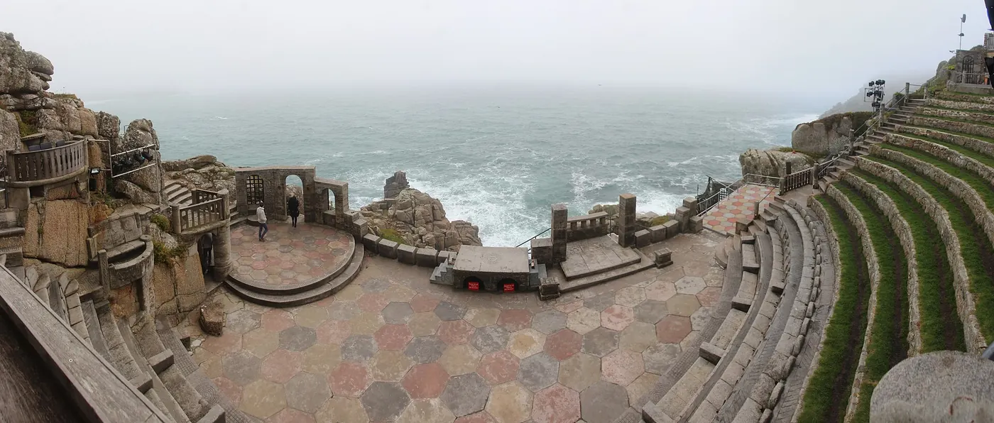 Wide panorama of the Minack stage of coloured hexagonal paving with the grey sea breaking on rocks behind and grassy seat terraces at right