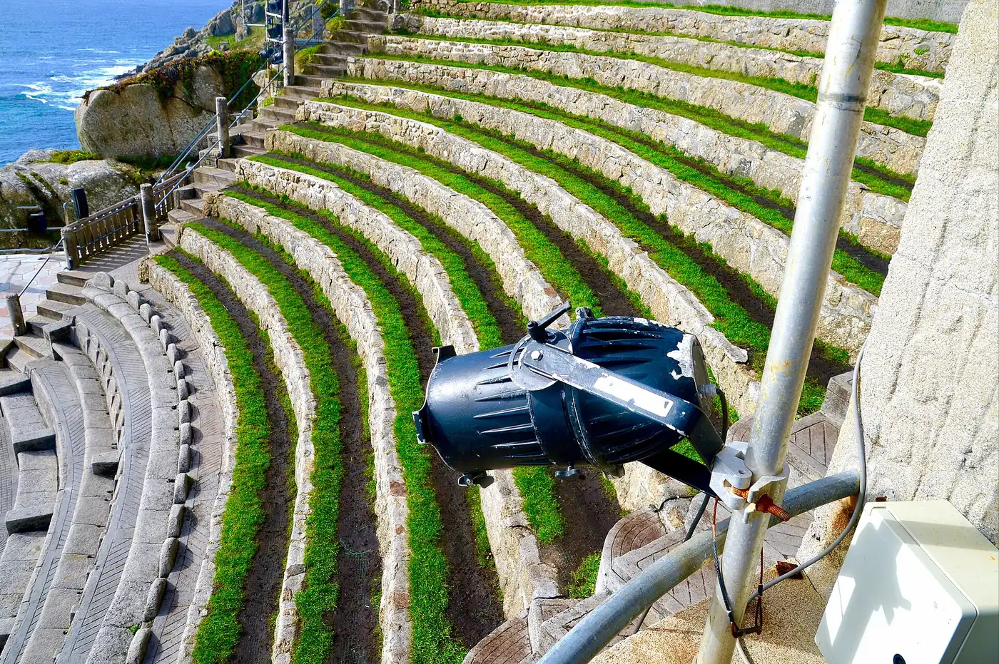 Curved grass-topped stone seating tiers at the Minack Theatre, with a stage light in the foreground and the sea in the top corner
