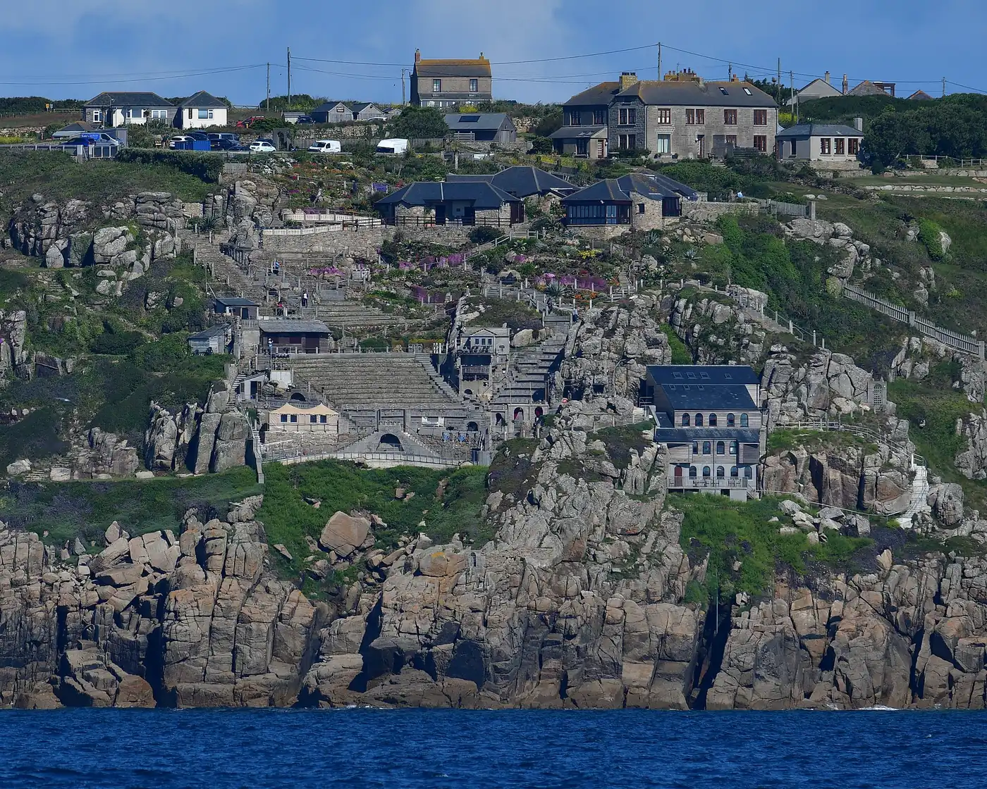 The Minack Theatre seen from the sea: stepped stone terraces, gardens and buildings climbing a granite cliff above rocks and blue water