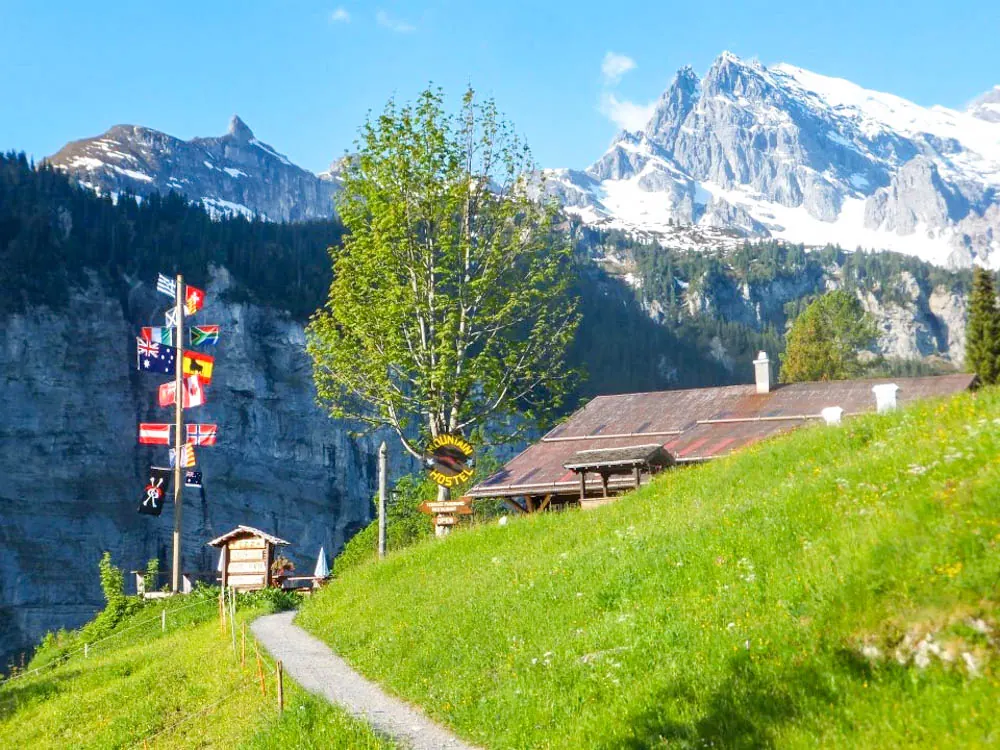 A green alpine slope and a chalet below snow-capped peaks