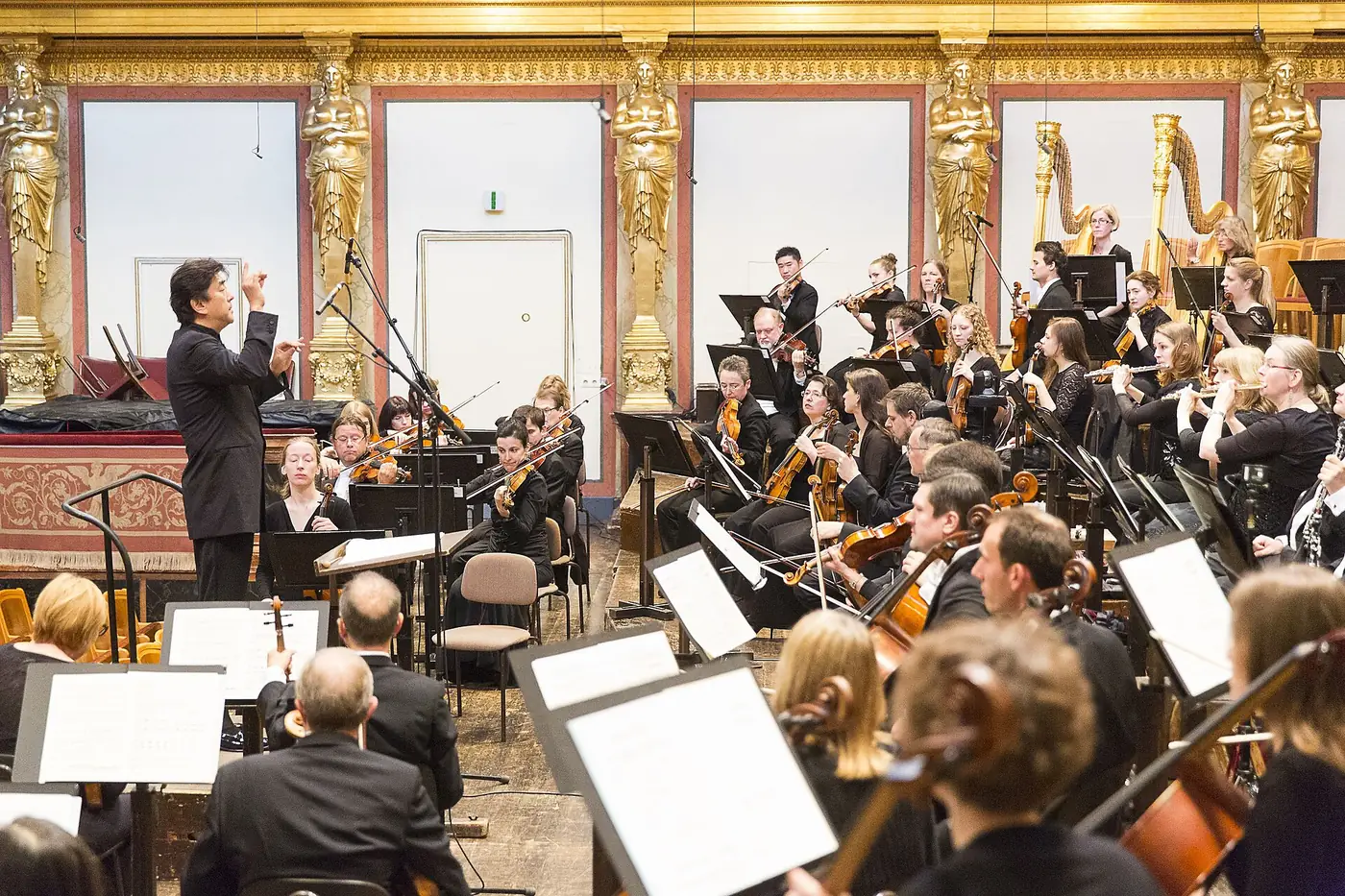 A conductor leads an orchestra on the Musikverein stage beneath gilded caryatids, with violins, flutes and harps in the foreground