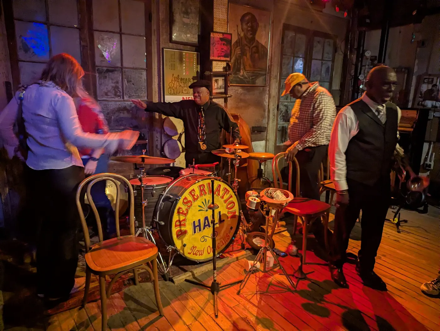 Musicians beside the painted Preservation Hall bass drum on the wooden stage in warm red light, a listener blurred at left