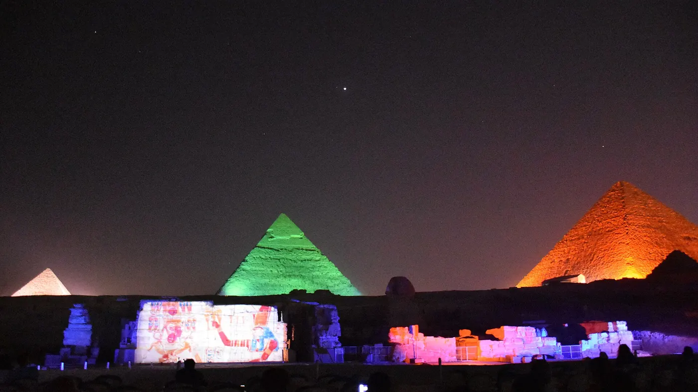 Pyramids lit green and orange above the Sphinx temple ruins, where an ancient painted scene is projected on the wall