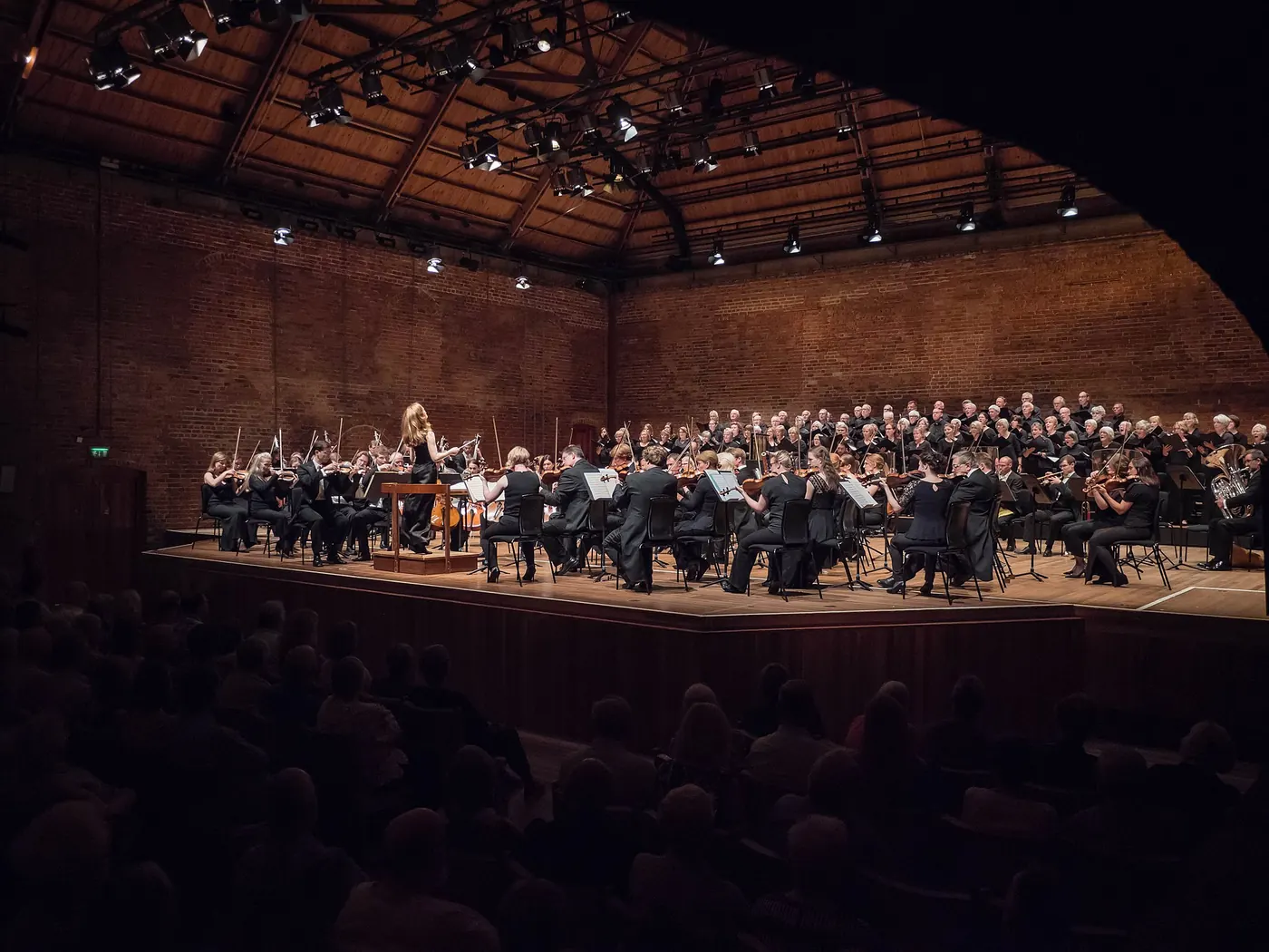 An orchestra and choir performing under a conductor at Snape Maltings, warm light on the brick walls and timber roof above the audience