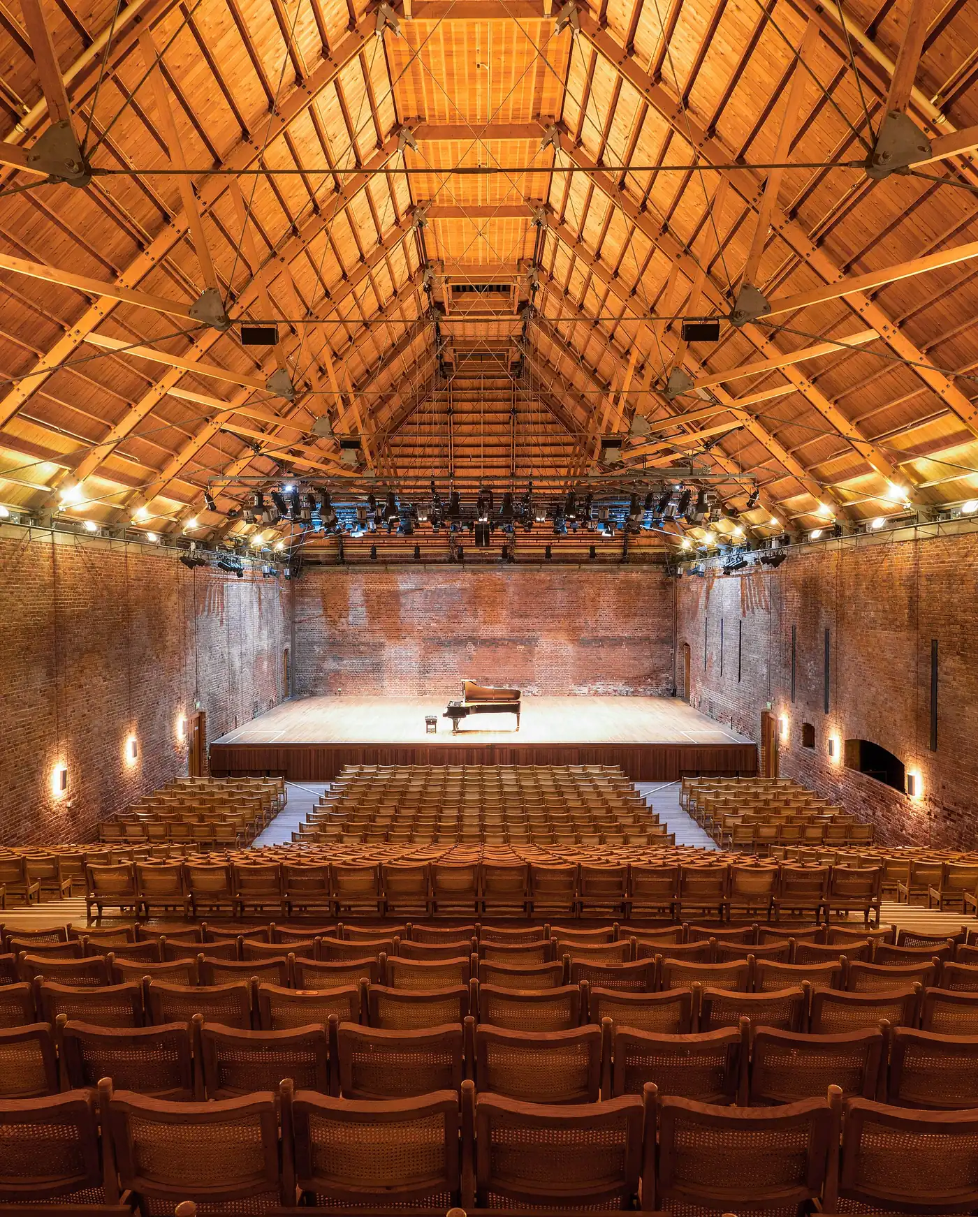 Empty Snape Maltings Concert Hall from the back: a grand piano on stage, rows of cane-backed chairs, bare brick walls and a timber roof