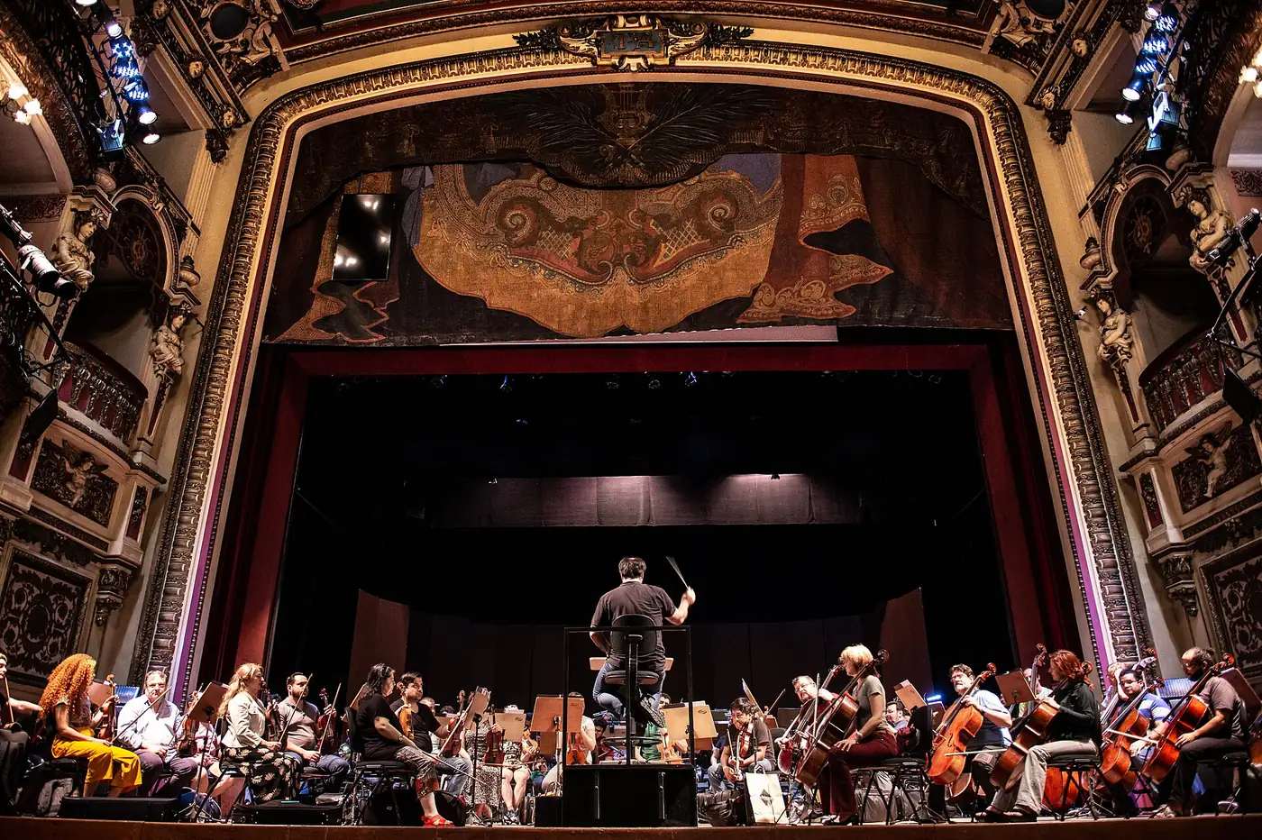 An orchestra rehearsing on the stage of Teatro Amazonas beneath the gilded proscenium arch and painted stage curtain