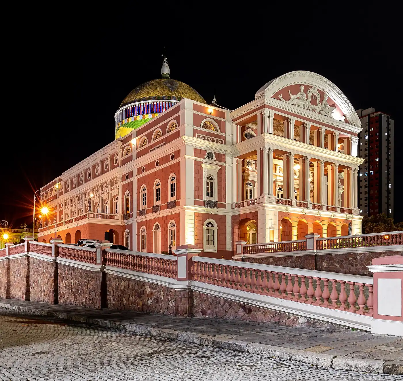 Teatro Amazonas at night, its pink and white facade floodlit beneath the tiled dome painted in the colours of Brazil's flag