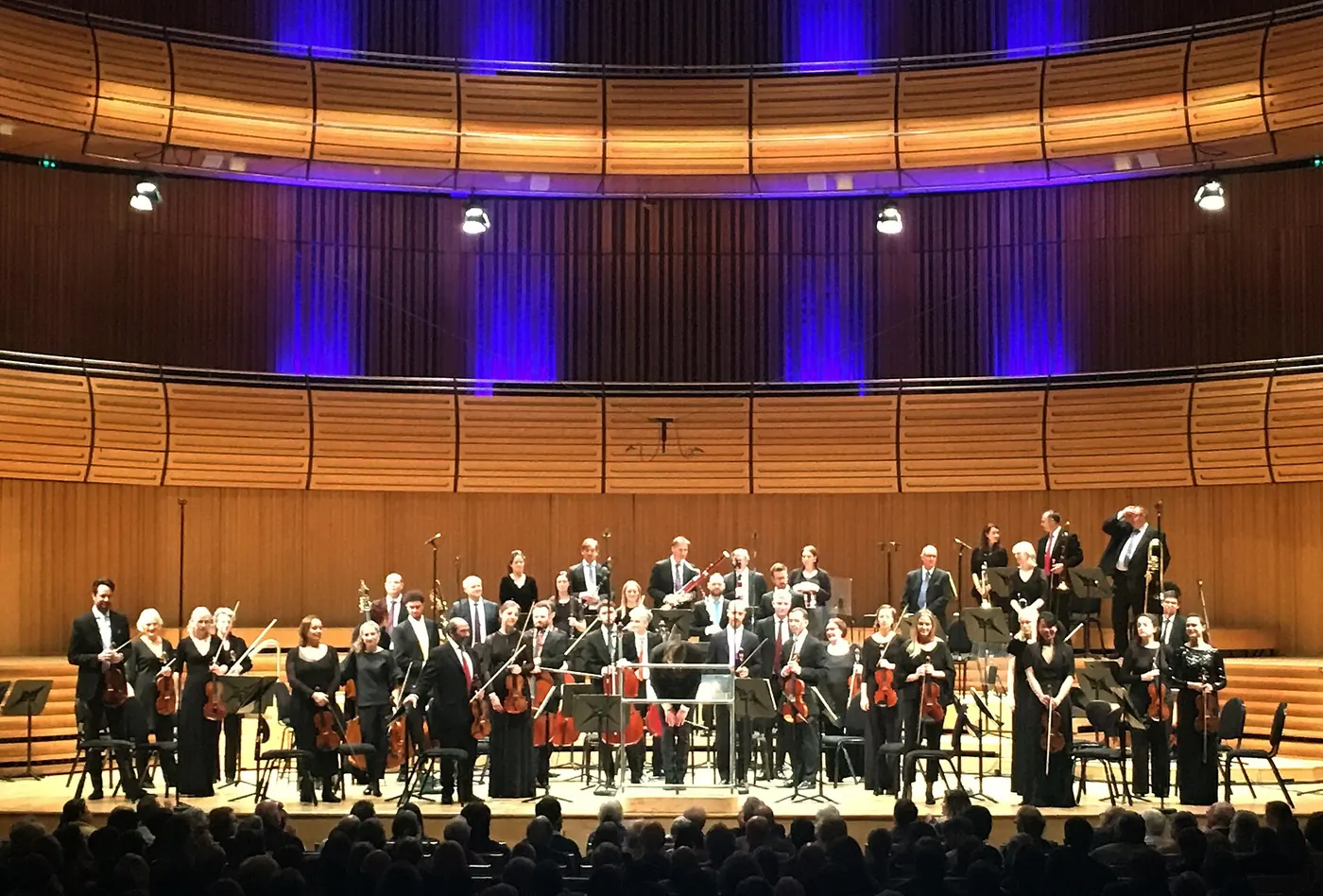 An orchestra stands to take applause on the wood-panelled stage of Hall One, with curved timber balconies lit in violet behind them