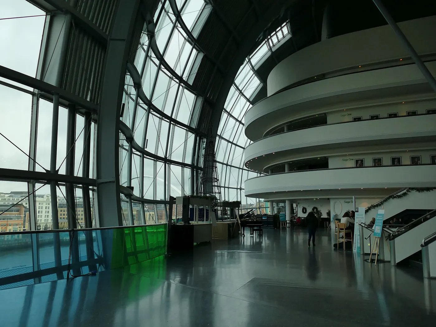 Inside the glass concourse of the Sage building, with tall curved glazing, city views and the white rings of Sage One beside a piano