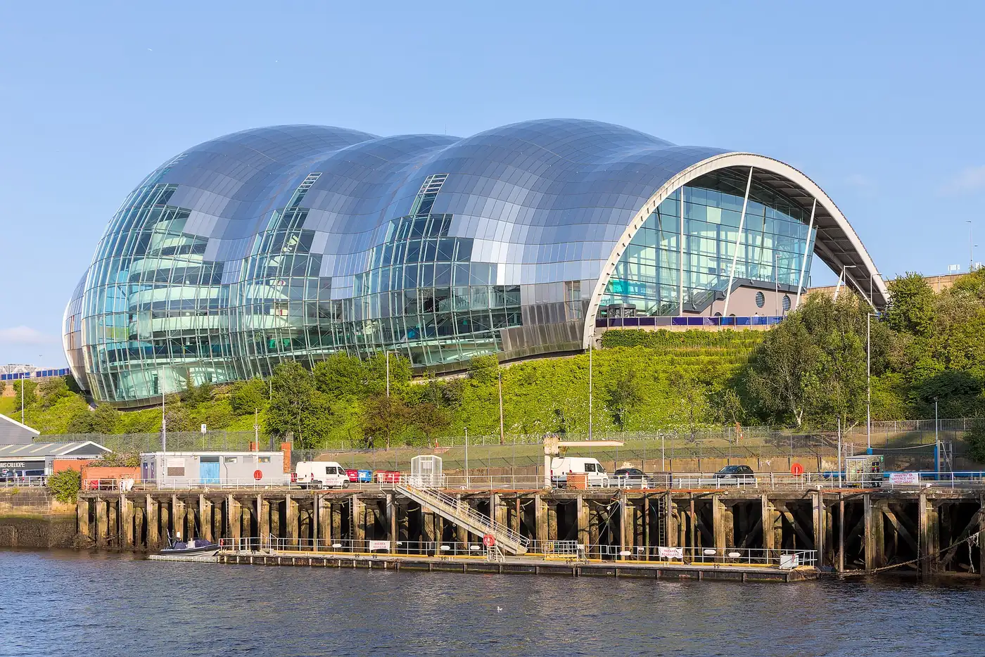 The Glasshouse in Gateshead seen from across the Tyne: a rippling steel-and-glass shell rising above grass banks and the old quayside