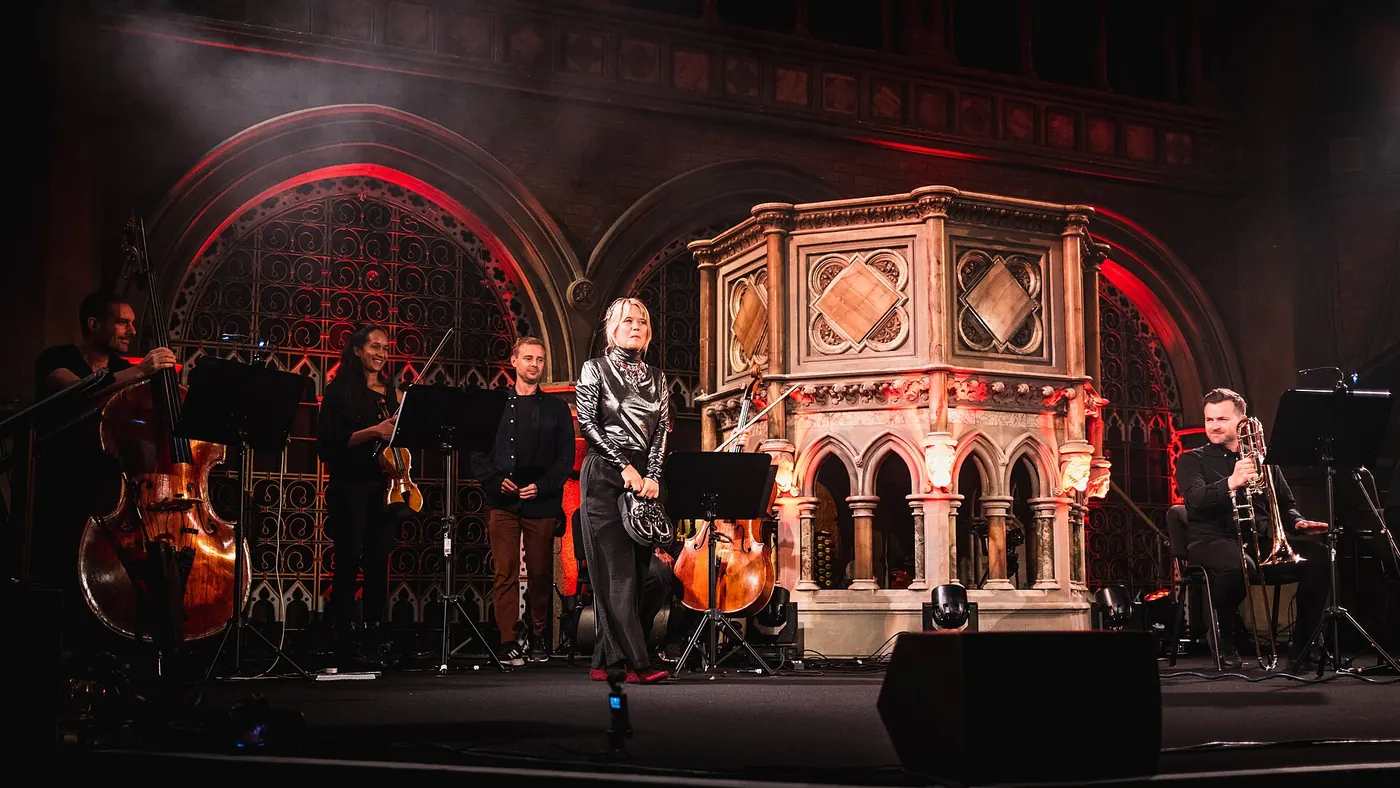 A band on stage at Union Chapel lit in red, in front of the carved stone pulpit and a Gothic iron screen, during Organ Reframed