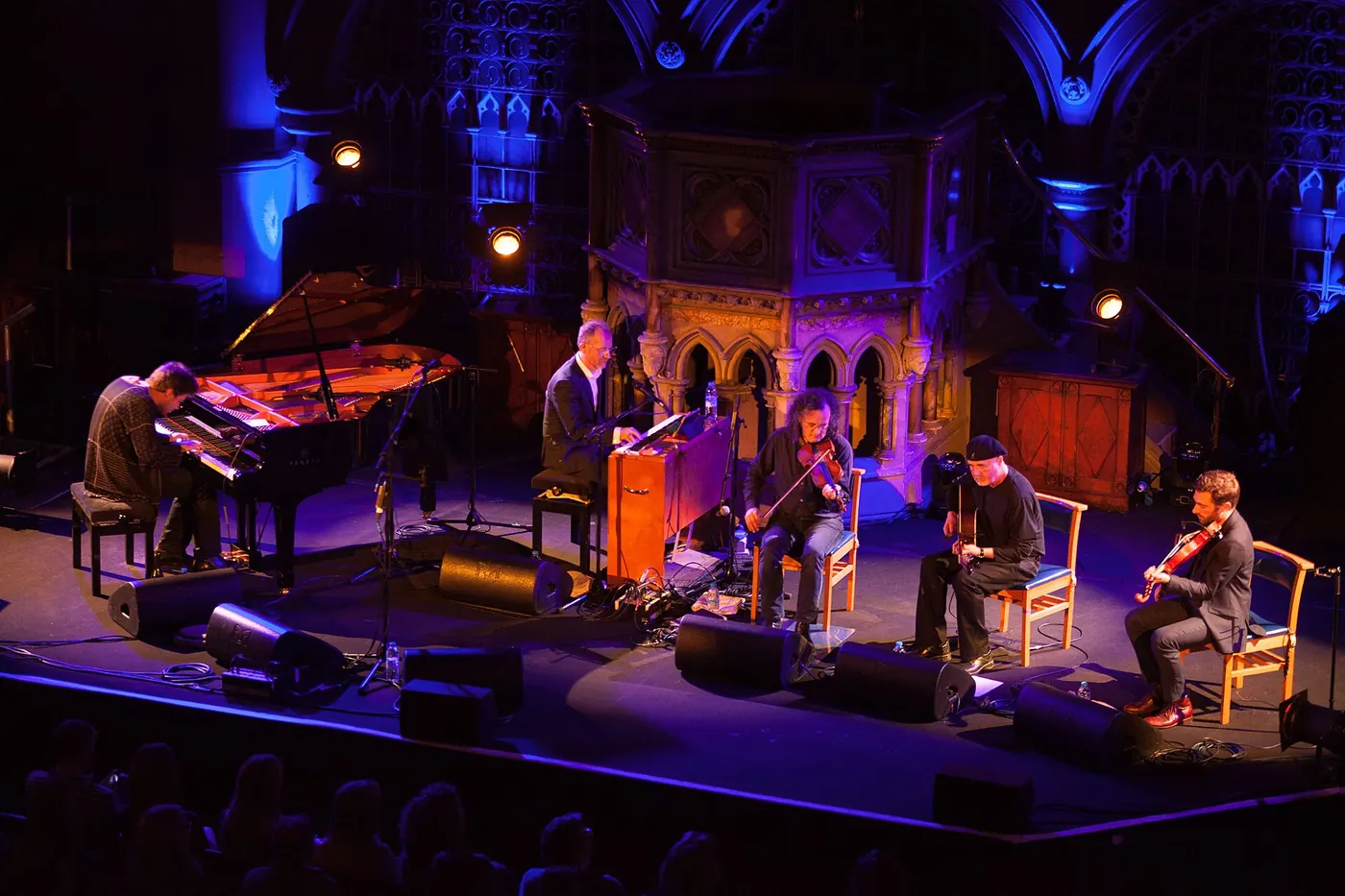 Five musicians on stage at Union Chapel, playing piano, fiddle and guitar in blue light before the stone pulpit, the audience silhouetted
