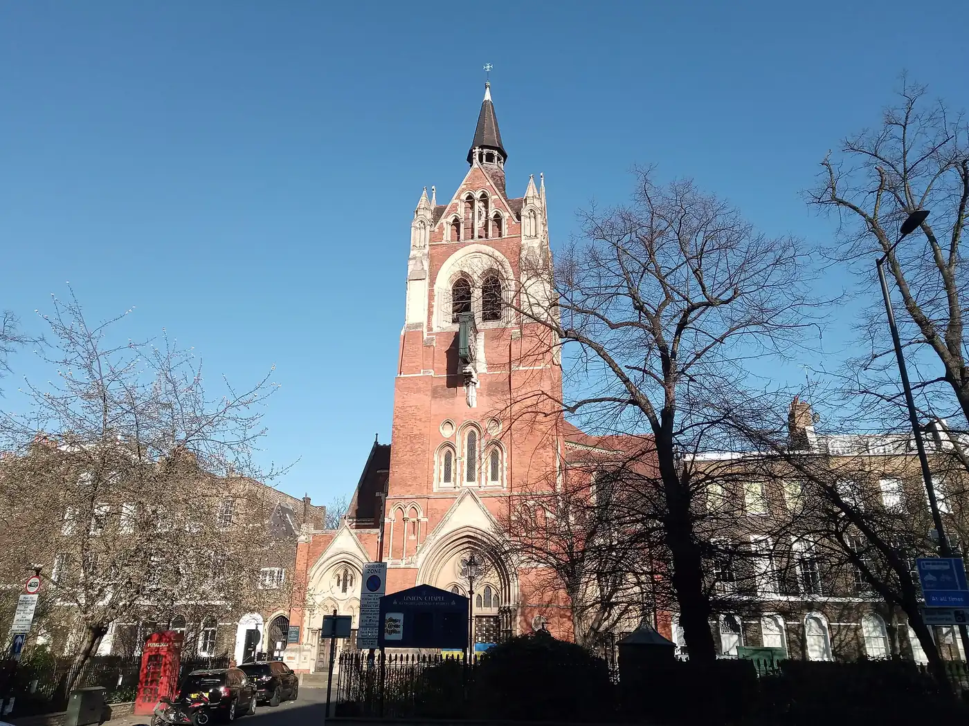 Union Chapel's red-brick tower and spire rising above the entrance on Compton Terrace, under a clear blue sky with bare trees in front
