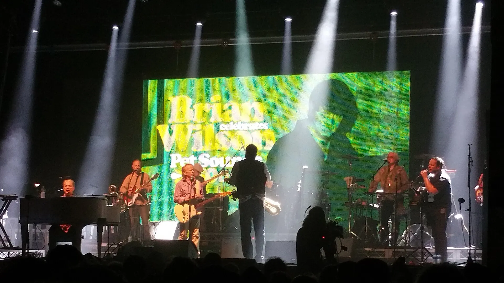A band on stage under bright lights in front of a screen reading Brian Wilson