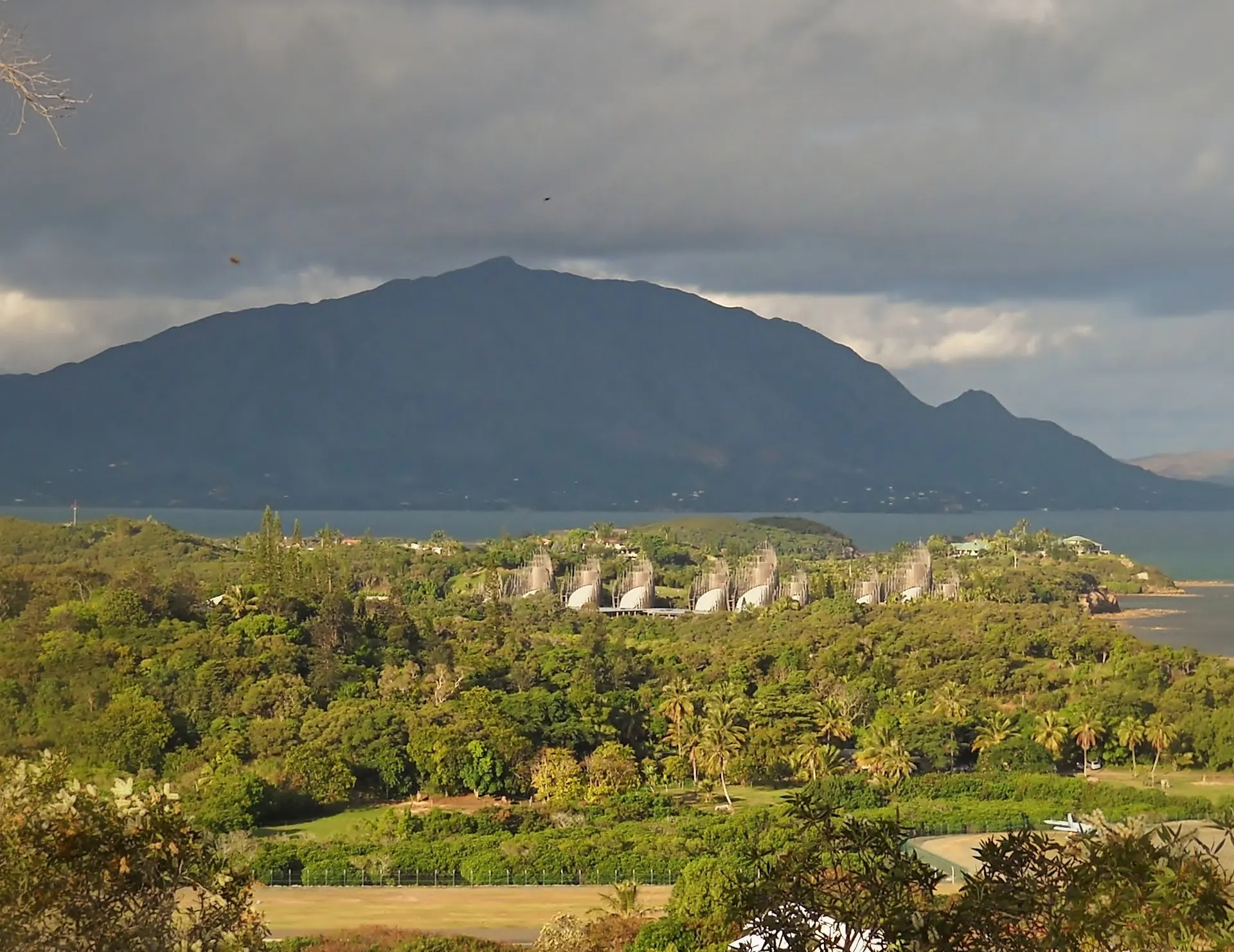The Centre Culturel Tjibaou seen across the bay: a row of tall timber huts among the trees, with the mountain behind
