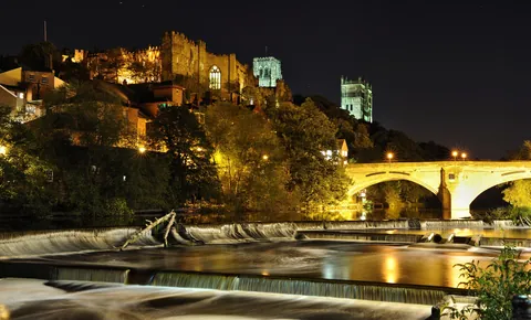 Durham Castle and Durham Cathedral lit at night above the River Wear, the weir and the bridge in the foreground