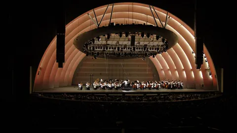 An orchestra in white jackets on the Hollywood Bowl stage, the glowing rings of the shell rising behind them into the night