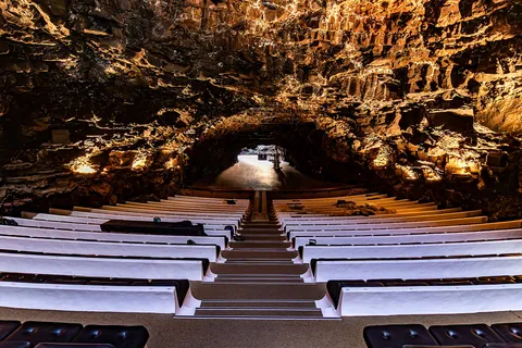Rows of white benches with black cushions descend through the lava tube auditorium towards a stage lit against rough basalt walls