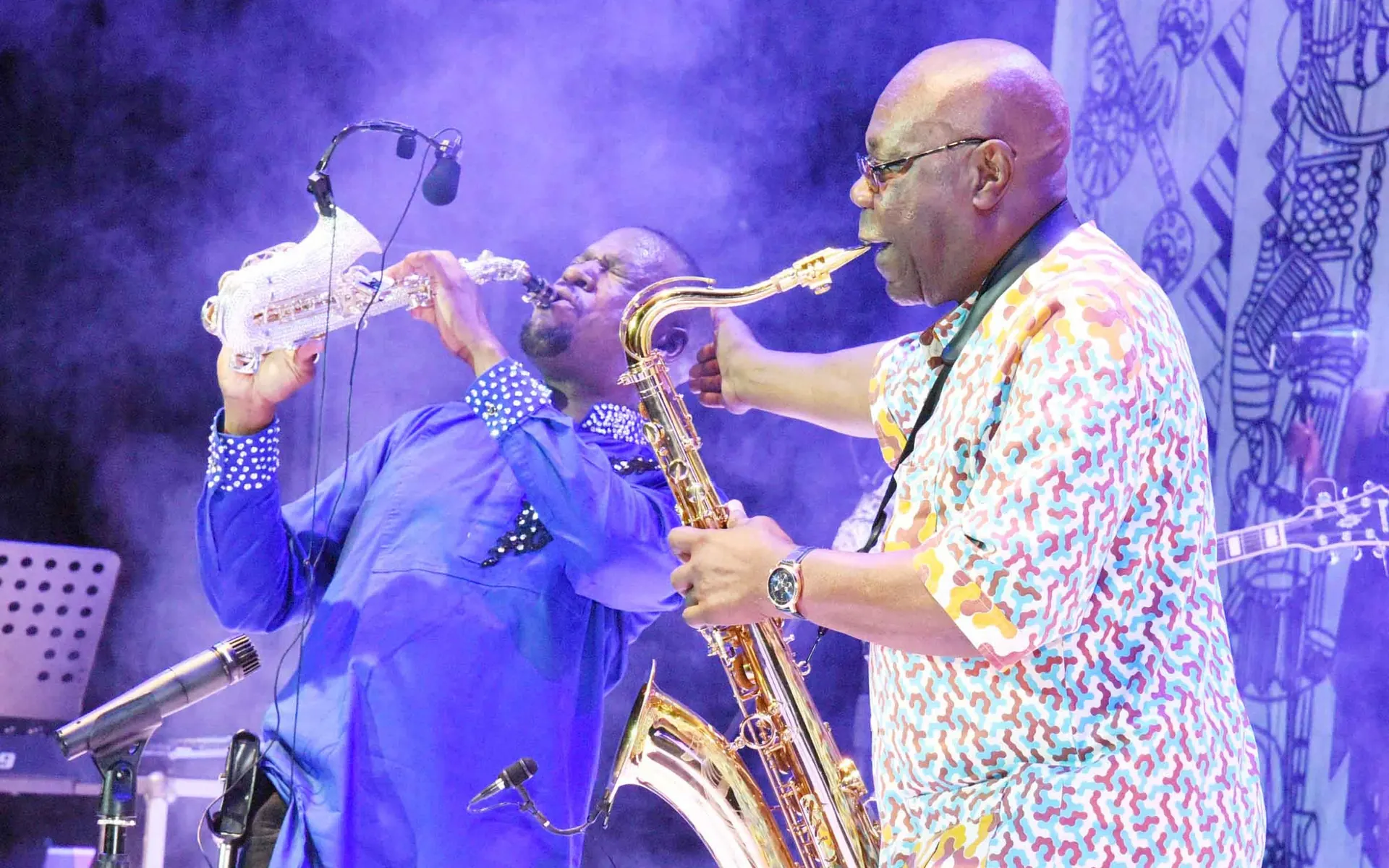 Two saxophonists playing together on stage, one in blue and one in a patterned shirt