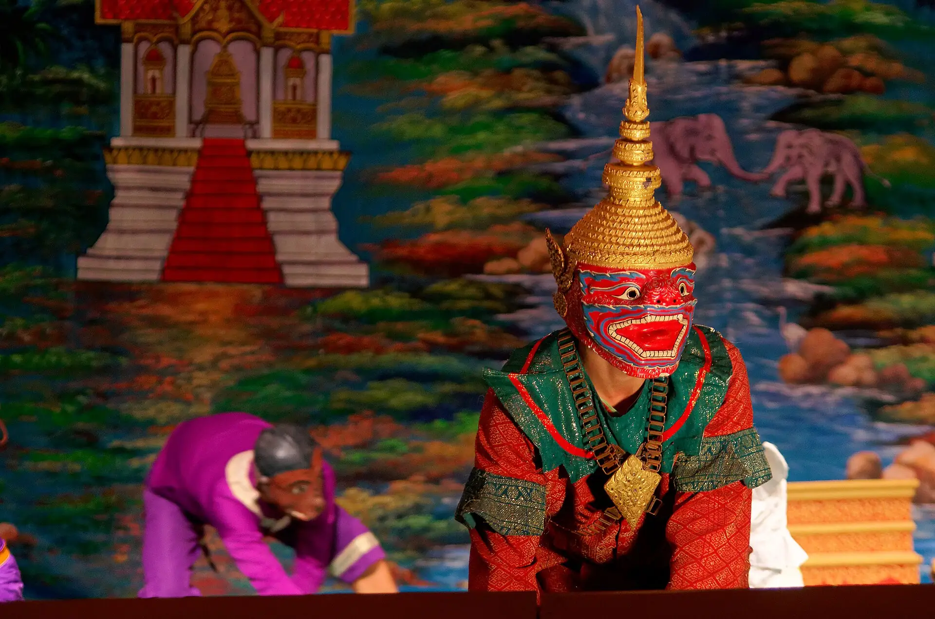 A performer in a red-and-green demon mask on a painted stage, from the Royal Ballet of Laos