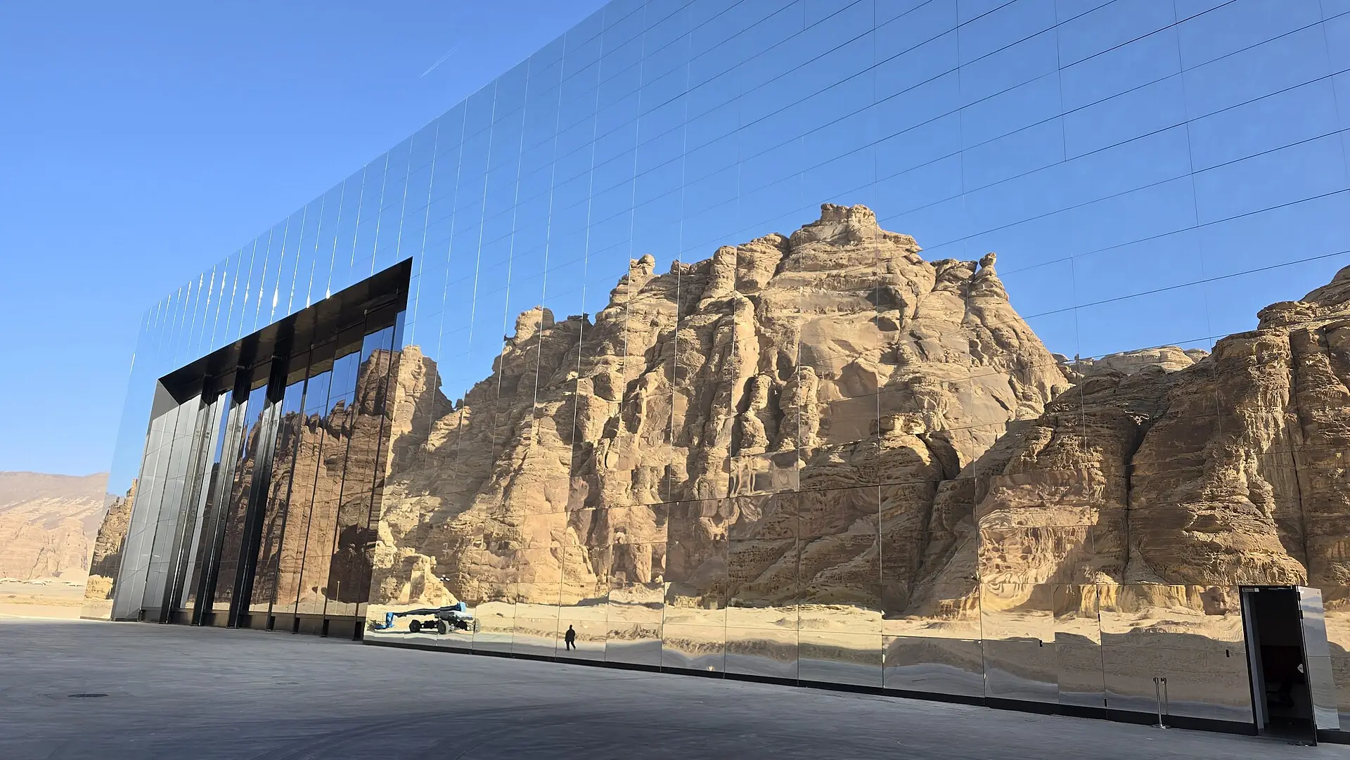 Close view of Maraya's mirror panels reflecting a towering sandstone crag and sky, with a tiny figure walking in the reflection