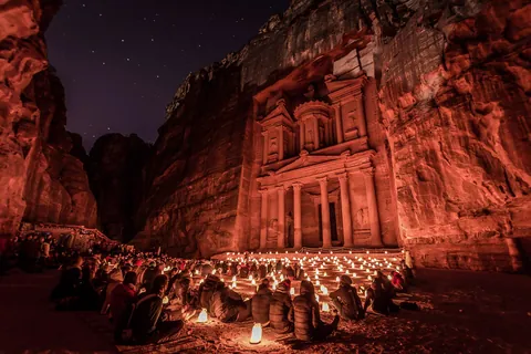 Crowd seated among candle lanterns in front of the Treasury at Petra, its carved facade glowing red under a starry night sky