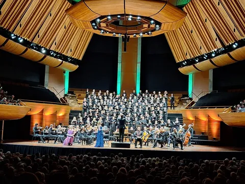 A choir and orchestra on stage in the Michael Fowler Centre, under a wooden ceiling