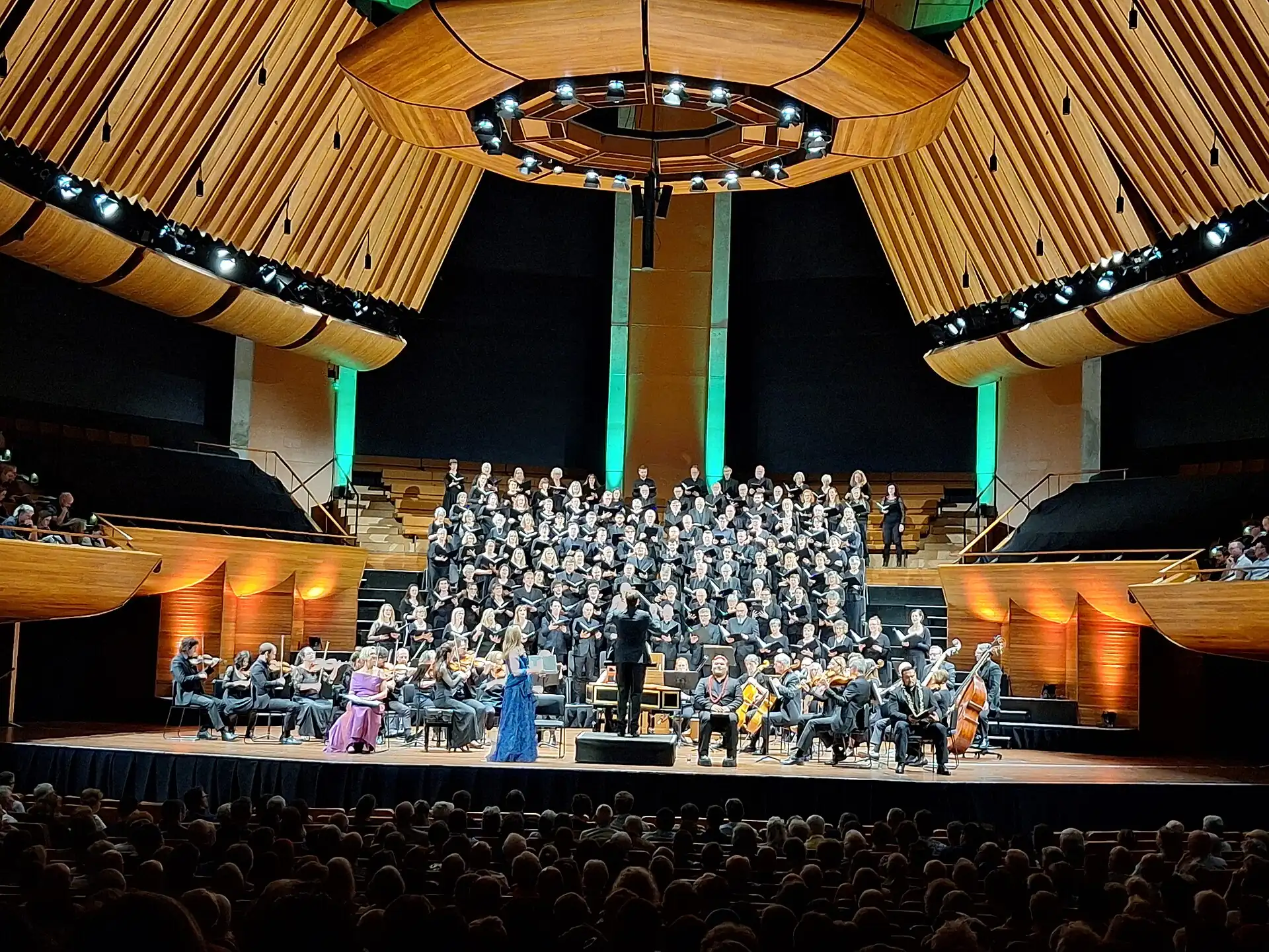 A choir and orchestra on stage in the Michael Fowler Centre, under a wooden ceiling