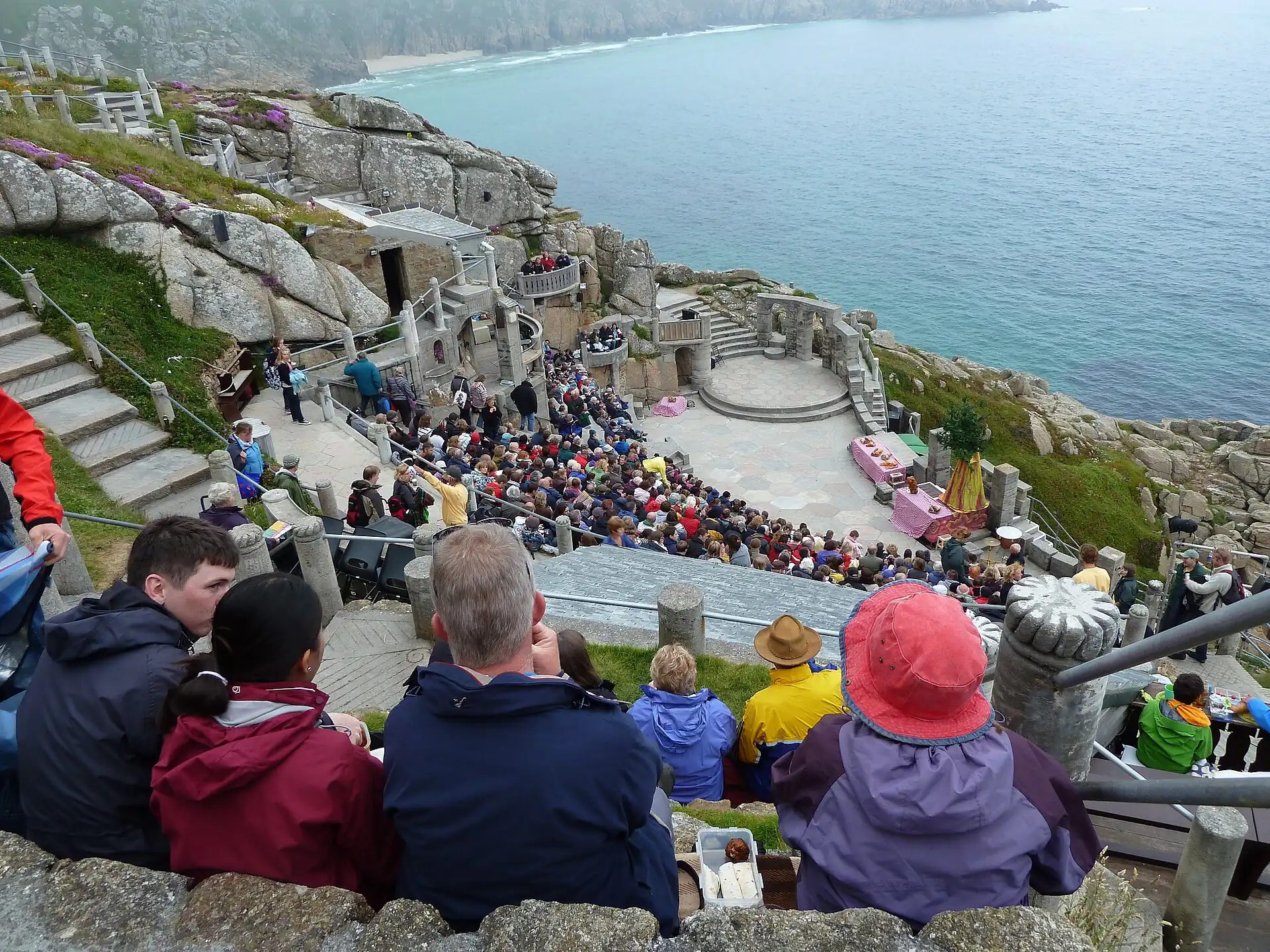 Audience seated in waterproofs on the stone terraces of the Minack during a matinee, the stage circle and turquoise sea beyond