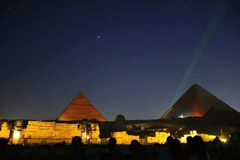 Audience silhouettes facing the Sphinx enclosure lit gold, with two pyramids beyond and a green laser under a starry sky