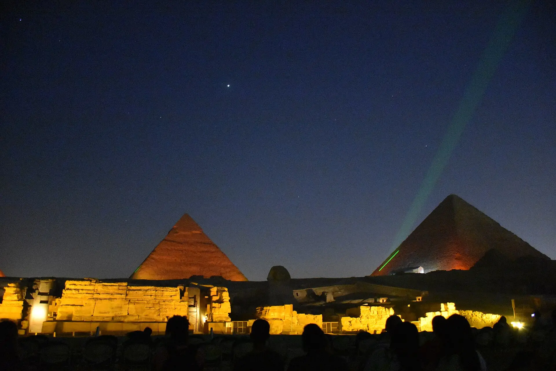 Audience silhouettes facing the Sphinx enclosure lit gold, with two pyramids beyond and a green laser under a starry sky