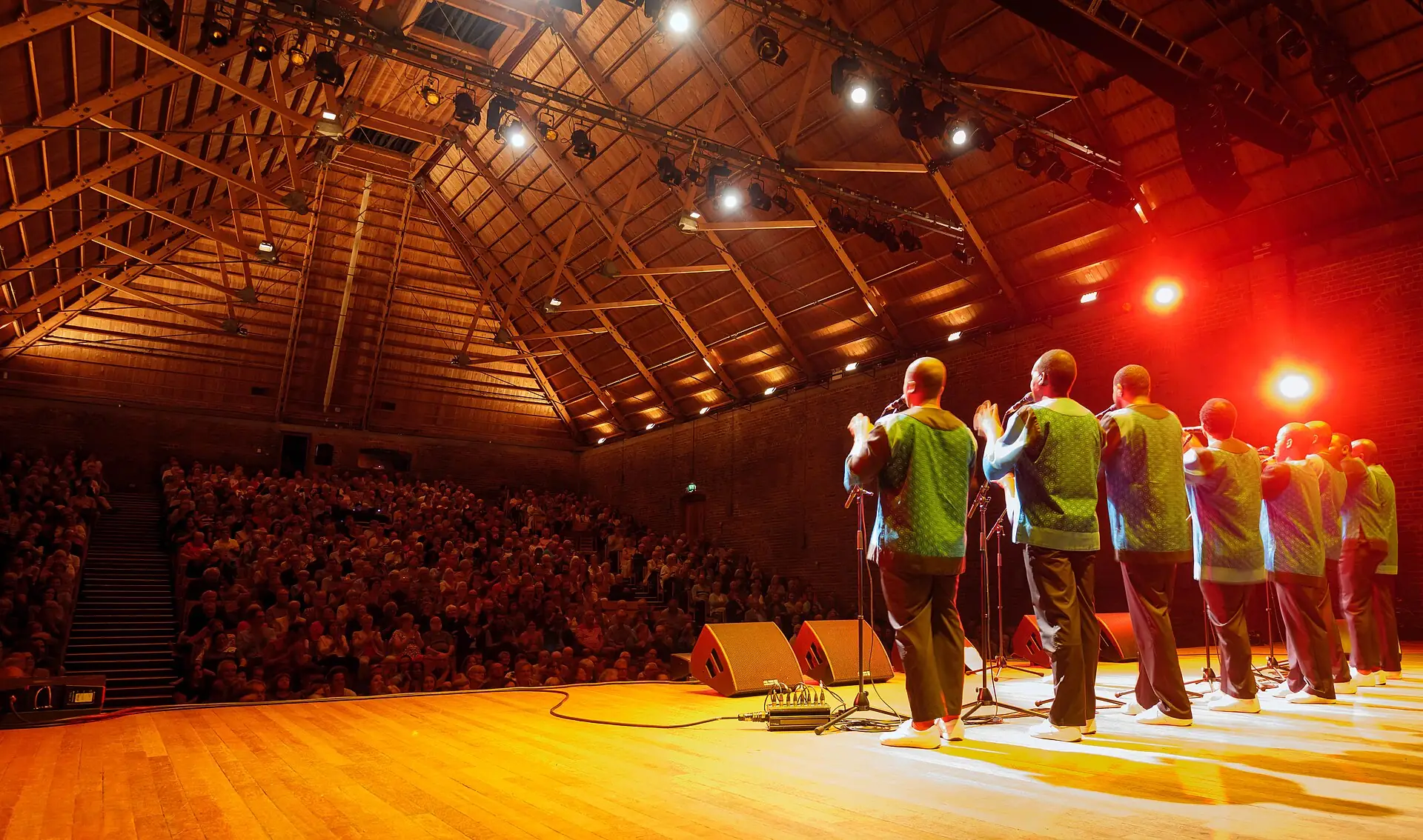 A vocal group performs on the stage of Snape Maltings Concert Hall, facing a full house under the timber roof trusses