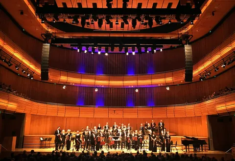 Wide view of Hall One's honey-coloured timber balconies and stage lighting, an orchestra taking a bow before a seated audience