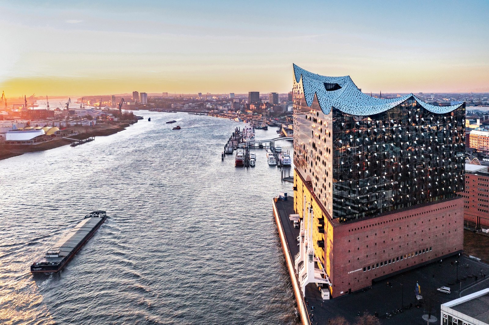 The Elbphilharmonie from above the Elbe at golden hour, its glass crown catching the light, a barge on the river and the harbour stretching away